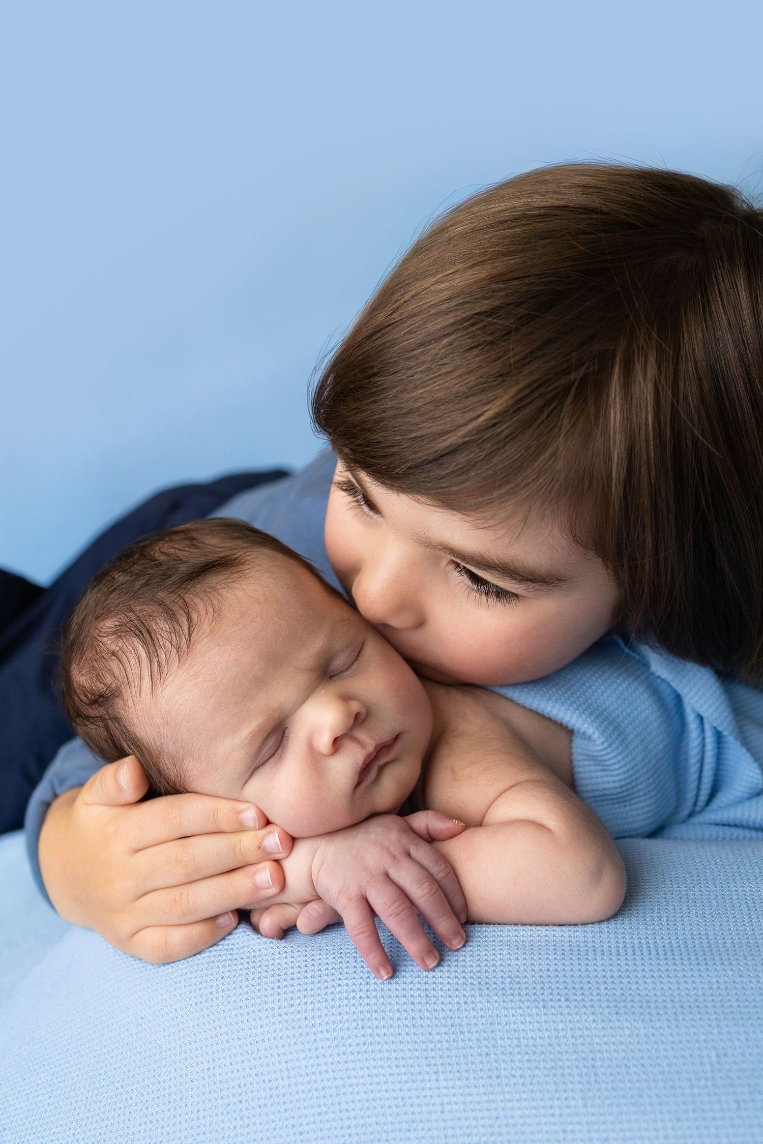 Older child in blue shirt gently kissing sleeping newborn baby on a soft blue background.