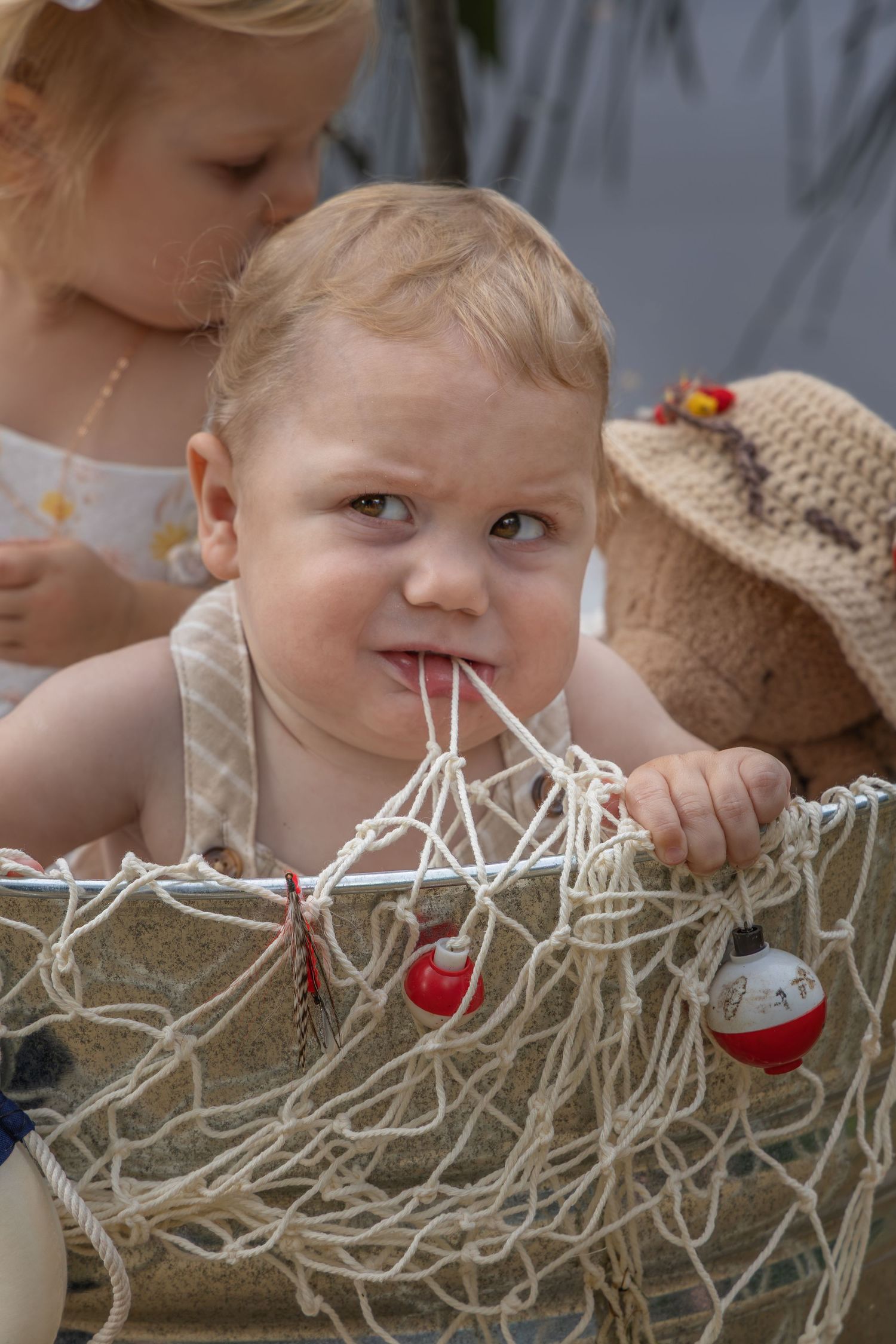 Curious baby chewing on fishing net rope with bobbers, sitting with older child and teddy bear.
