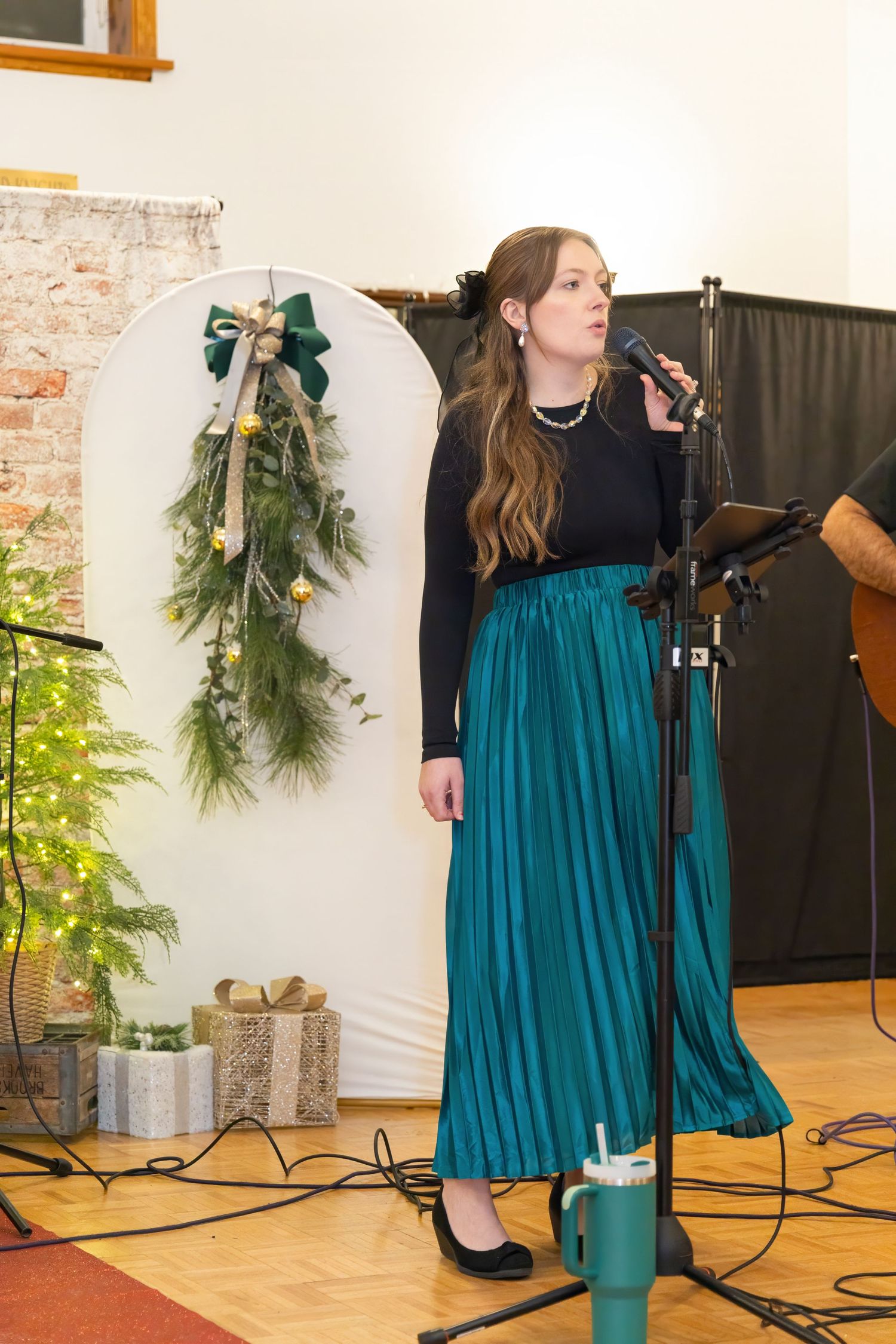 A performer in a teal dress sings on stage with holiday greenery decorations.