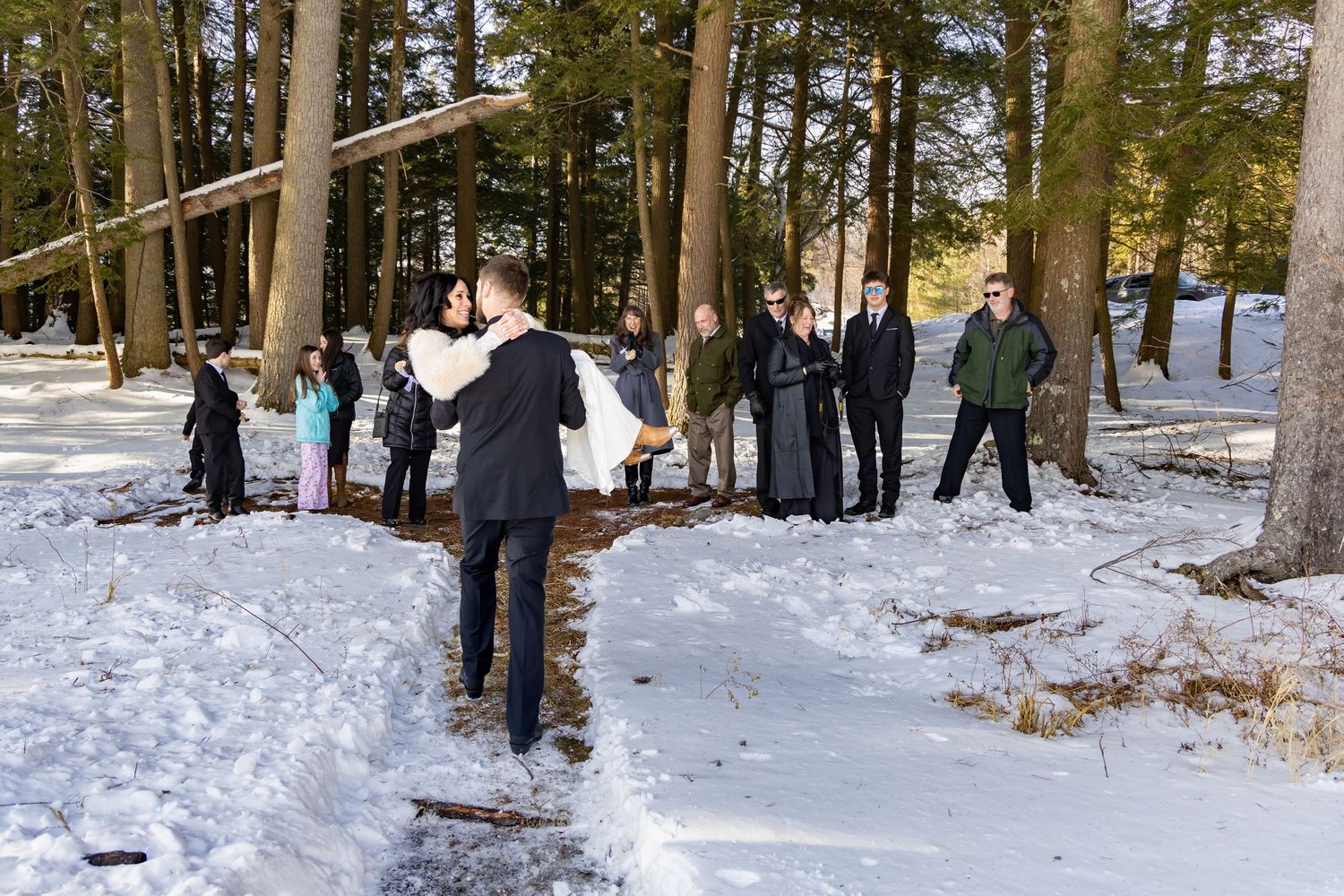 Winter wedding guests gather on a snow-covered path surrounded by evergreen trees.