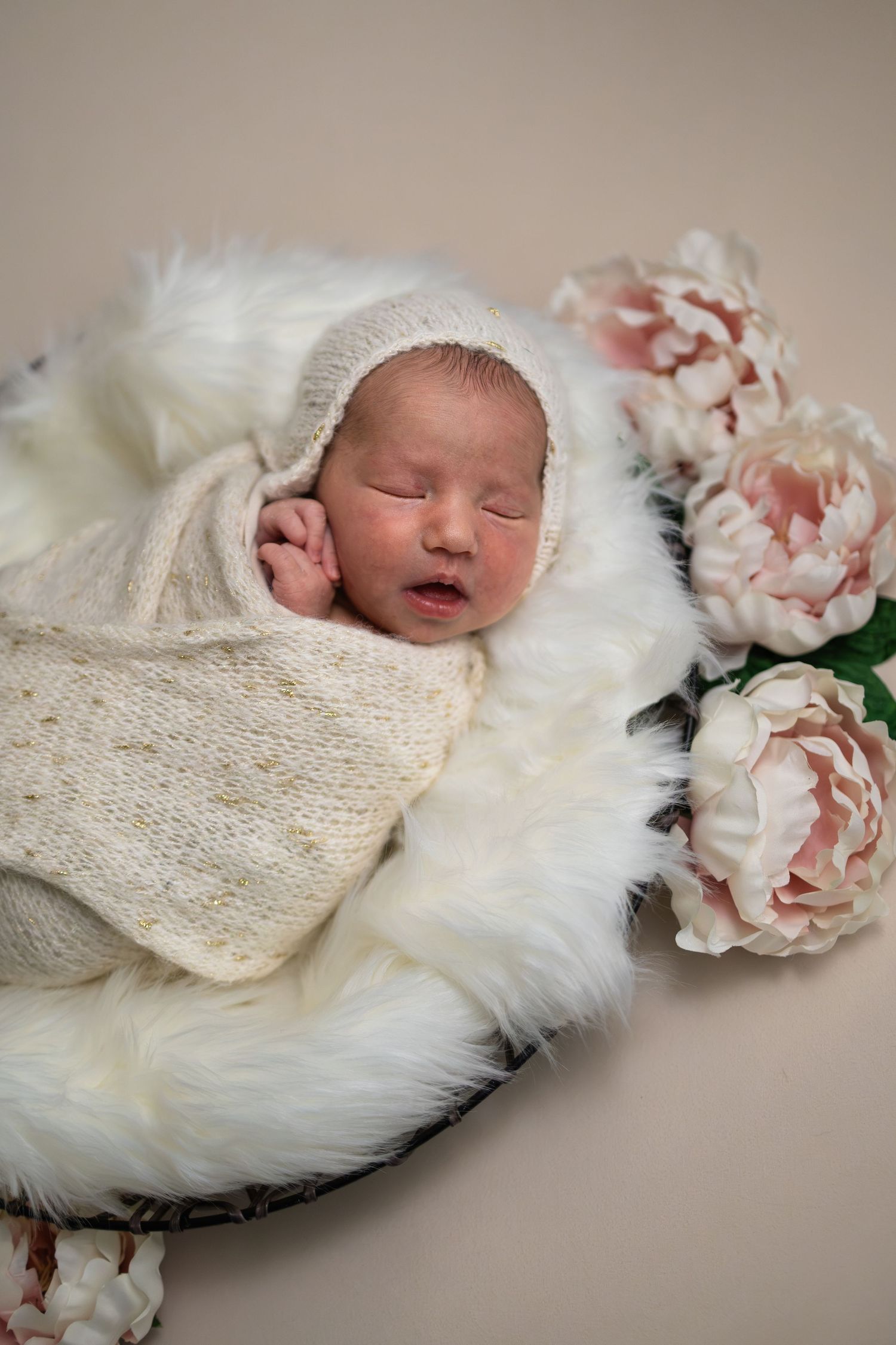 Newborn baby sleeping in a basket with white fur and pink peonies, wrapped in cream knit blanket and bonnet.