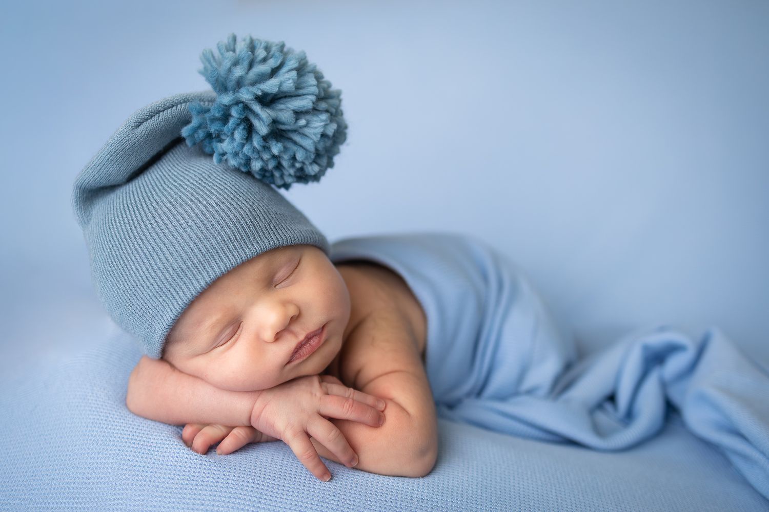 Sleeping newborn baby wearing a blue knit pom-pom hat, wrapped in a soft blue blanket on a blue background.