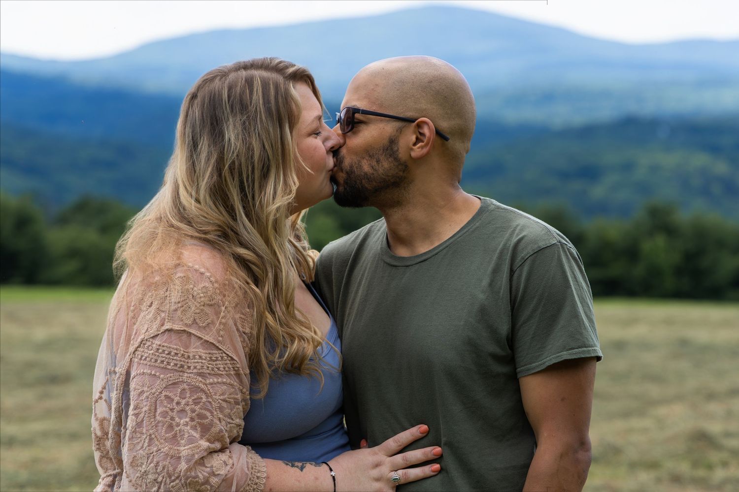Couple sharing a romantic kiss in a green field with scenic mountain views in the background.
