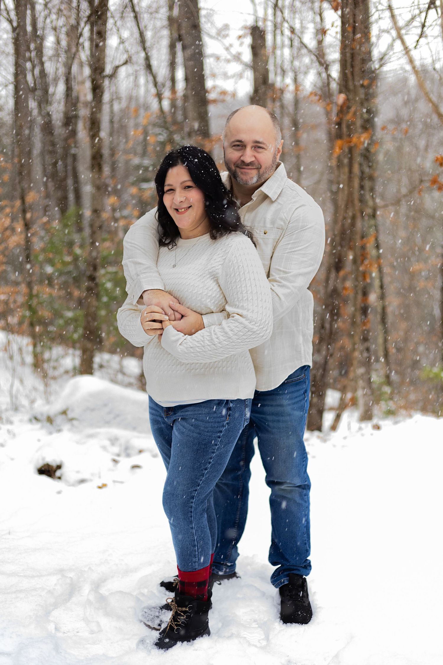A couple embraces in a snowy winter forest scene wearing matching cream sweaters and jeans.