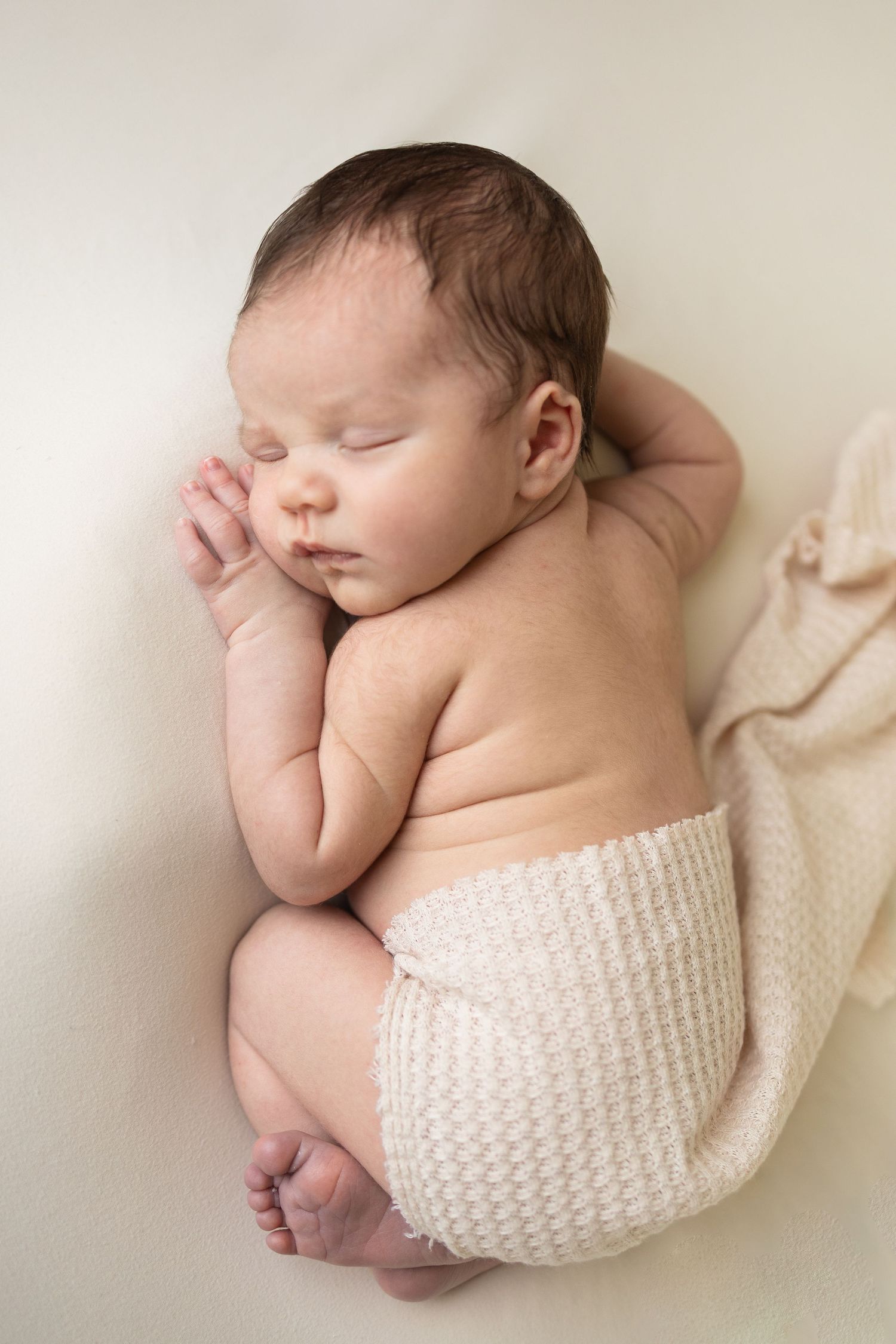 Peaceful newborn baby sleeping curled up on white blanket, wearing cream knit shorts, with dark hair and chubby cheeks.