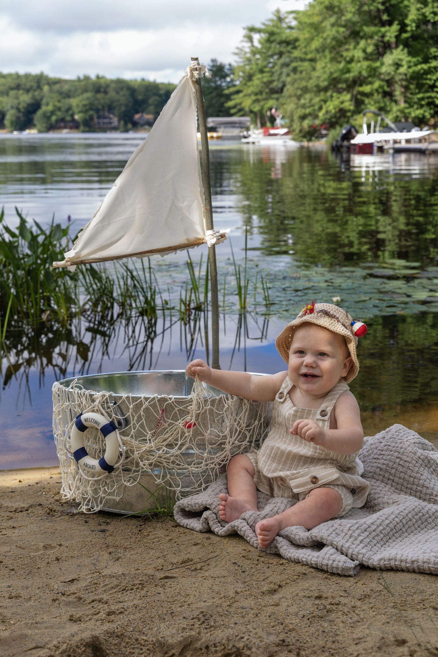 Happy baby in sailor hat sits on sandy beach beside decorative toy sailboat with lake and dock in background.