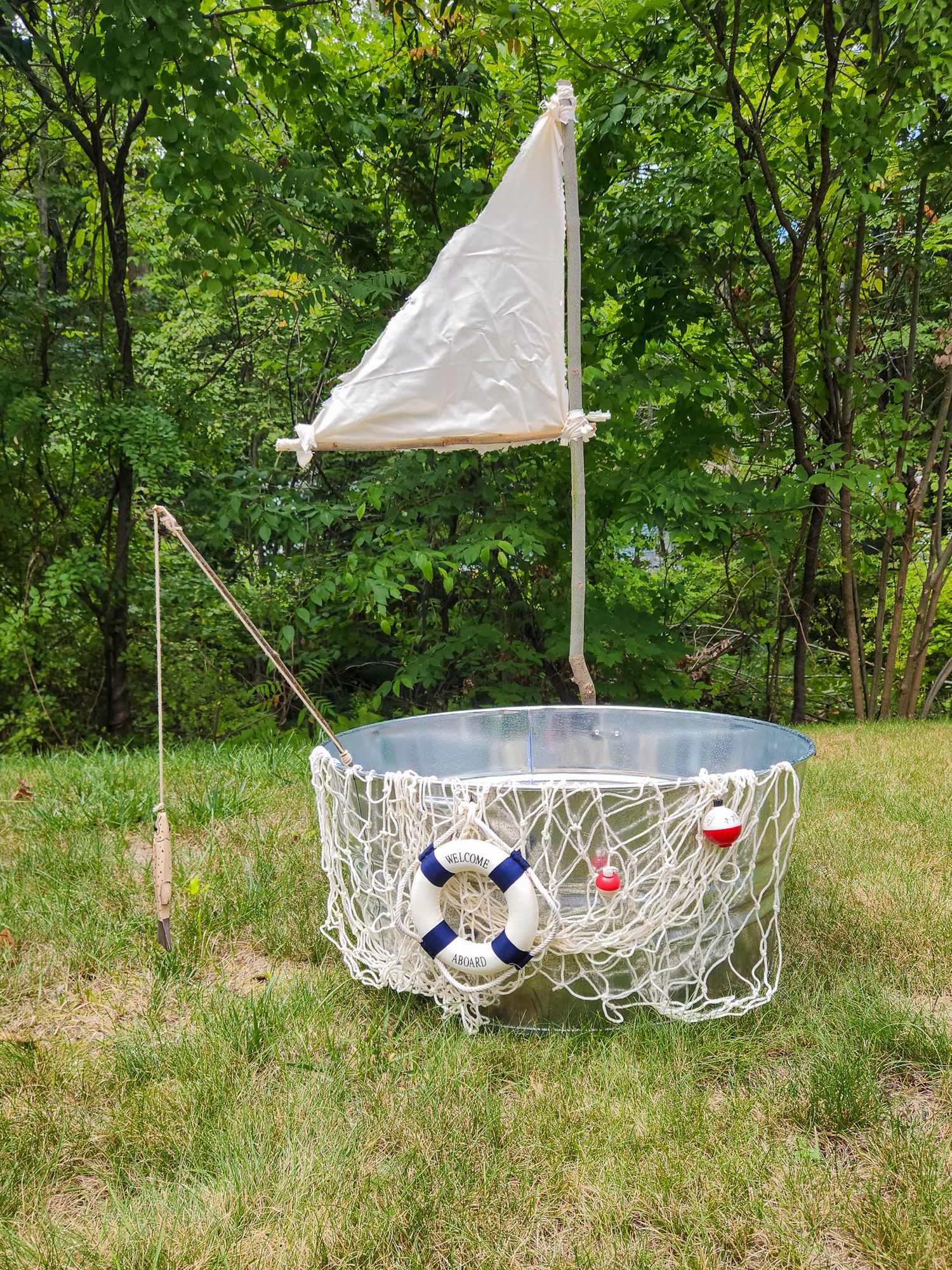 Decorative galvanized tub with nautical theme, featuring a sail, rope netting, and life preserver ring on green lawn.