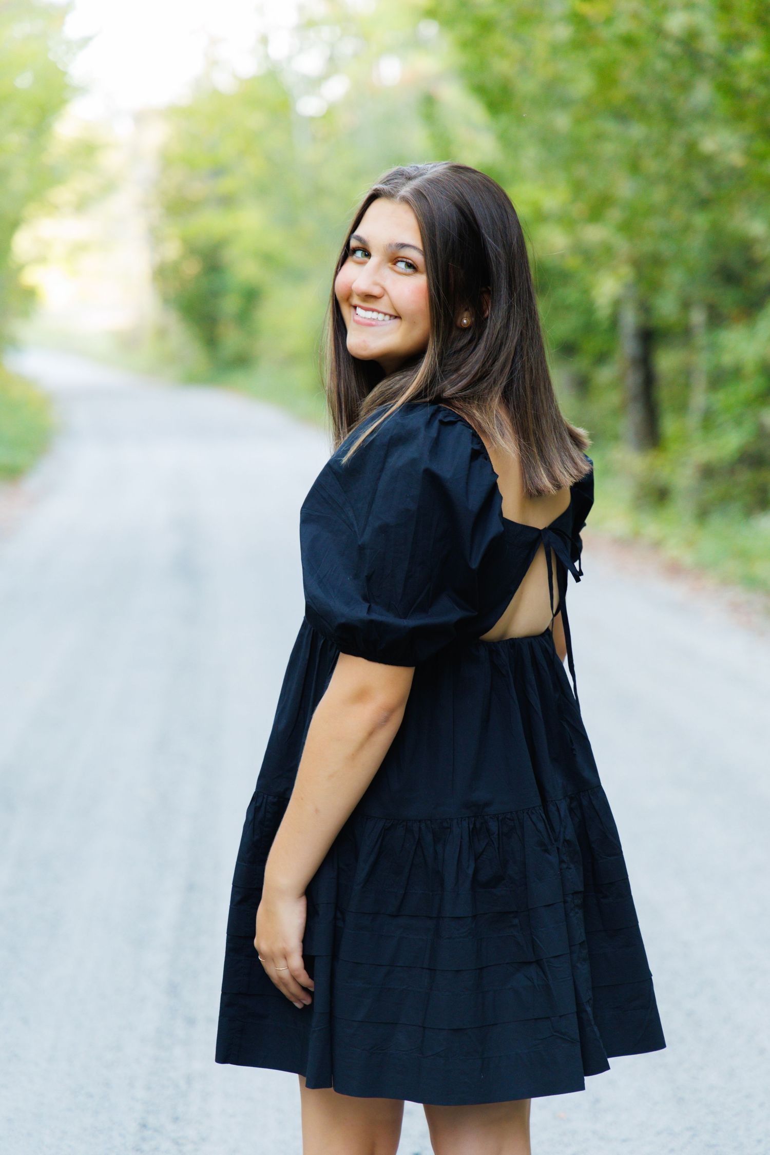 A person in a black dress twirls and poses playfully on a country road.