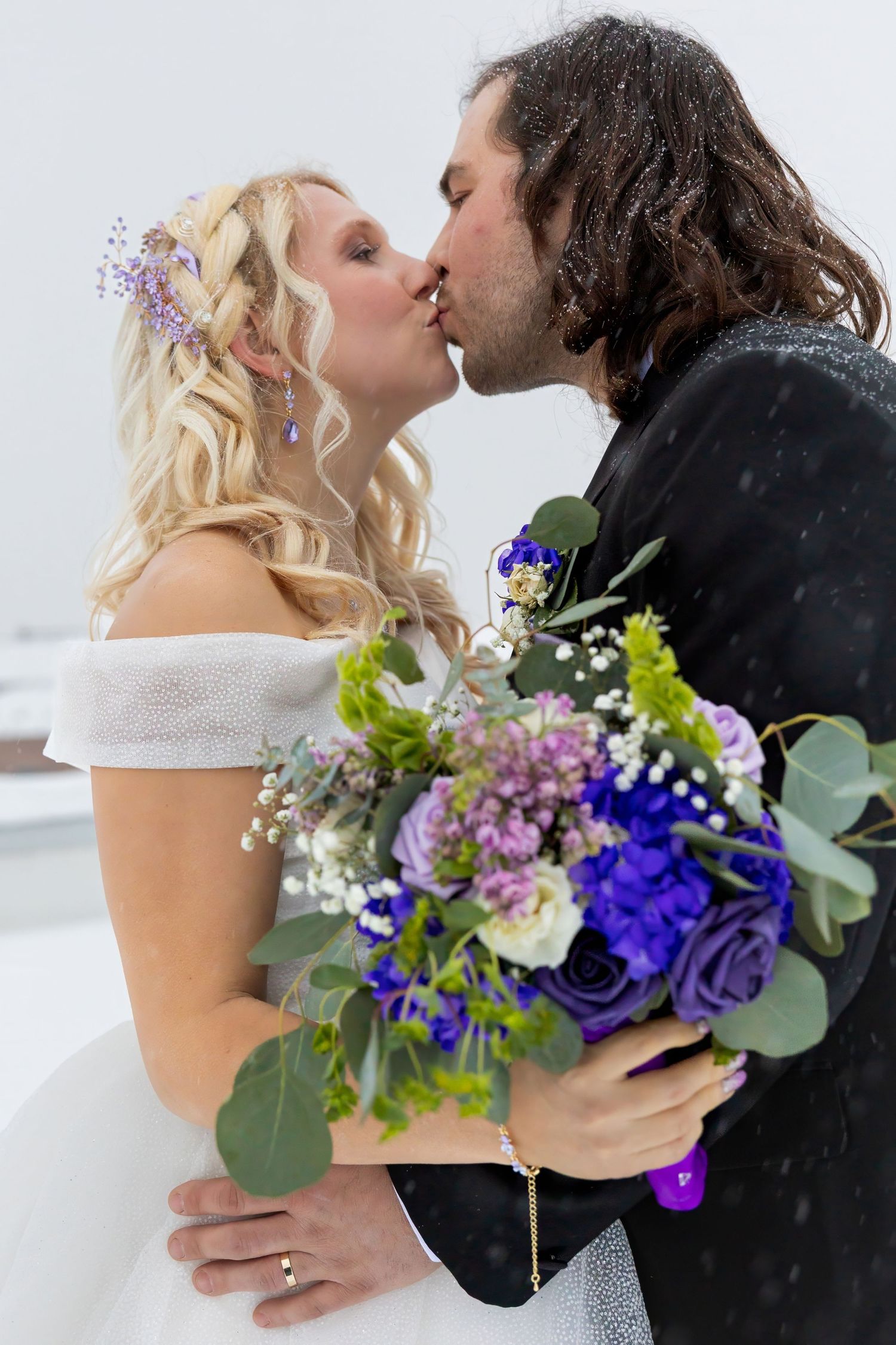 Winter wedding couple embraces with purple and blue floral bouquet against snowy backdrop.