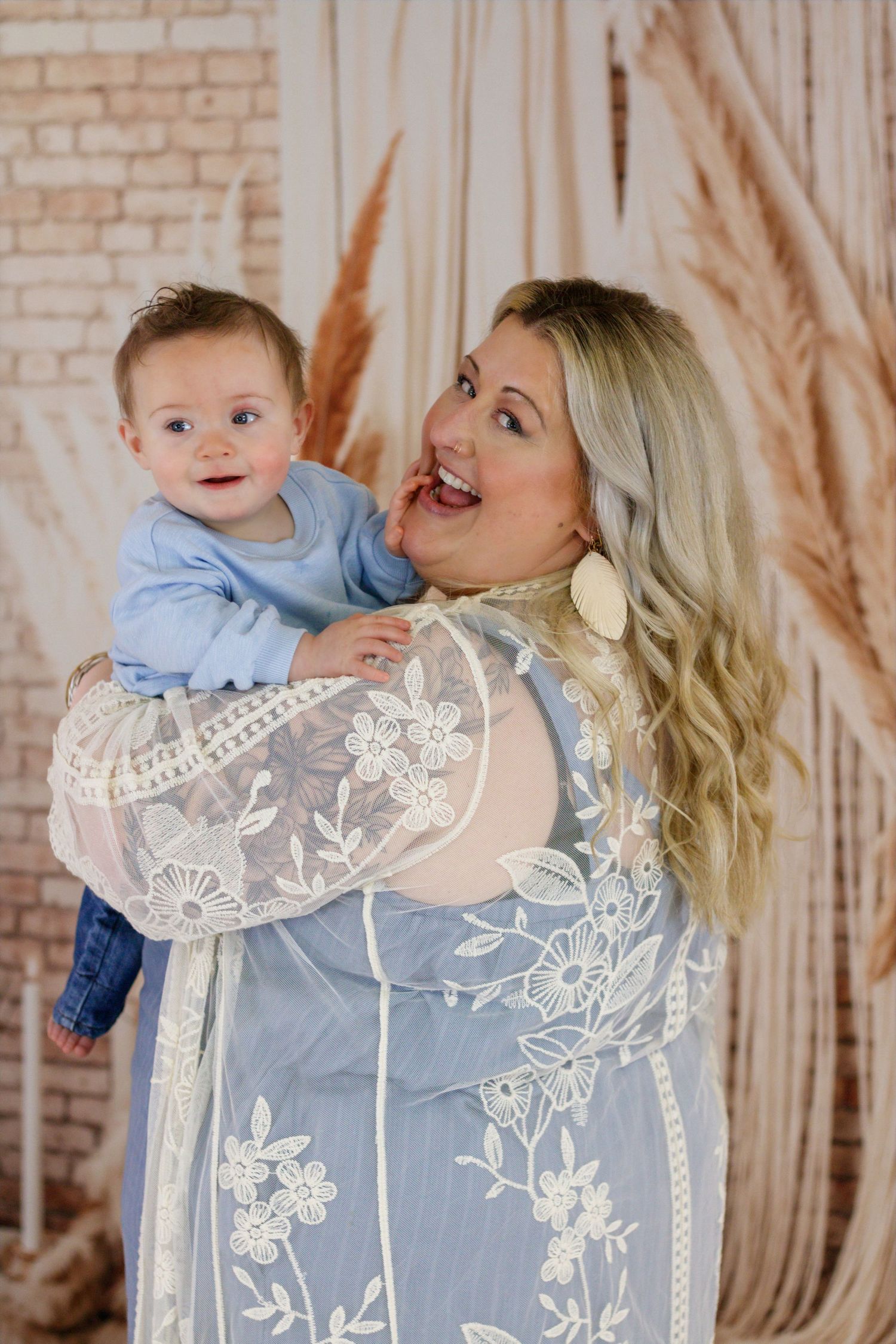 Smiling blonde woman in floral lace dress holding a happy baby boy in blue outfit against boho backdrop.