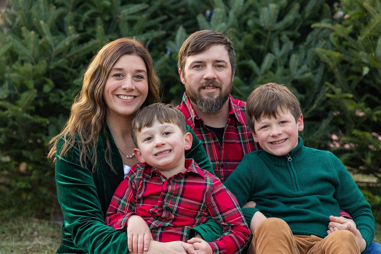 Family of four smiling outdoors at a Christmas tree farm, parents with two young boys in festive holiday attire.