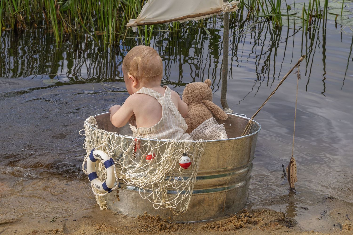 Baby sitting in a metal tub by a lake shore, with a toy sailboat and fishing rod, enjoying a playful outdoor adventure.