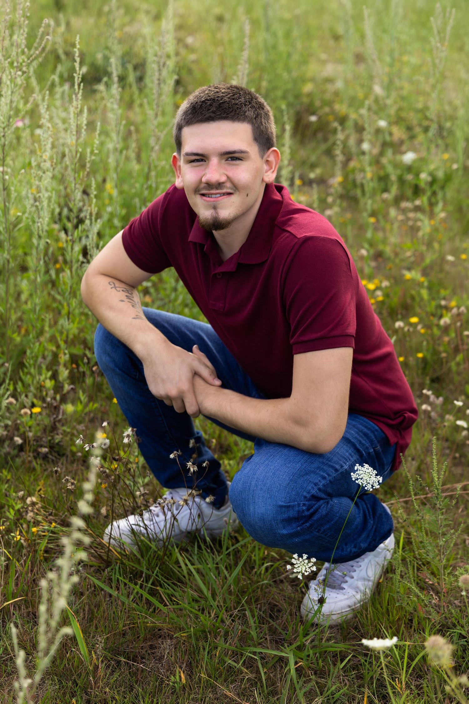 Young man in burgundy polo and jeans crouching in a wildflower meadow, smiling at the camera.