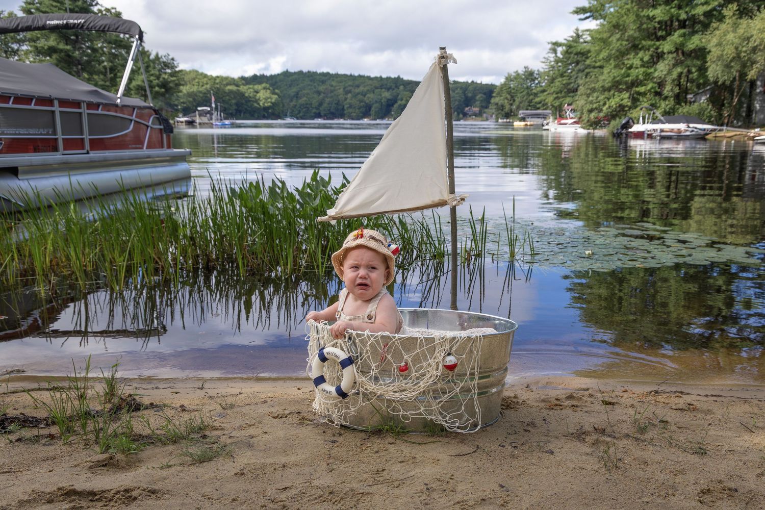 Baby in sailor hat sits in decorative wire boat basket on sandy lakeshore with boats and trees in background.