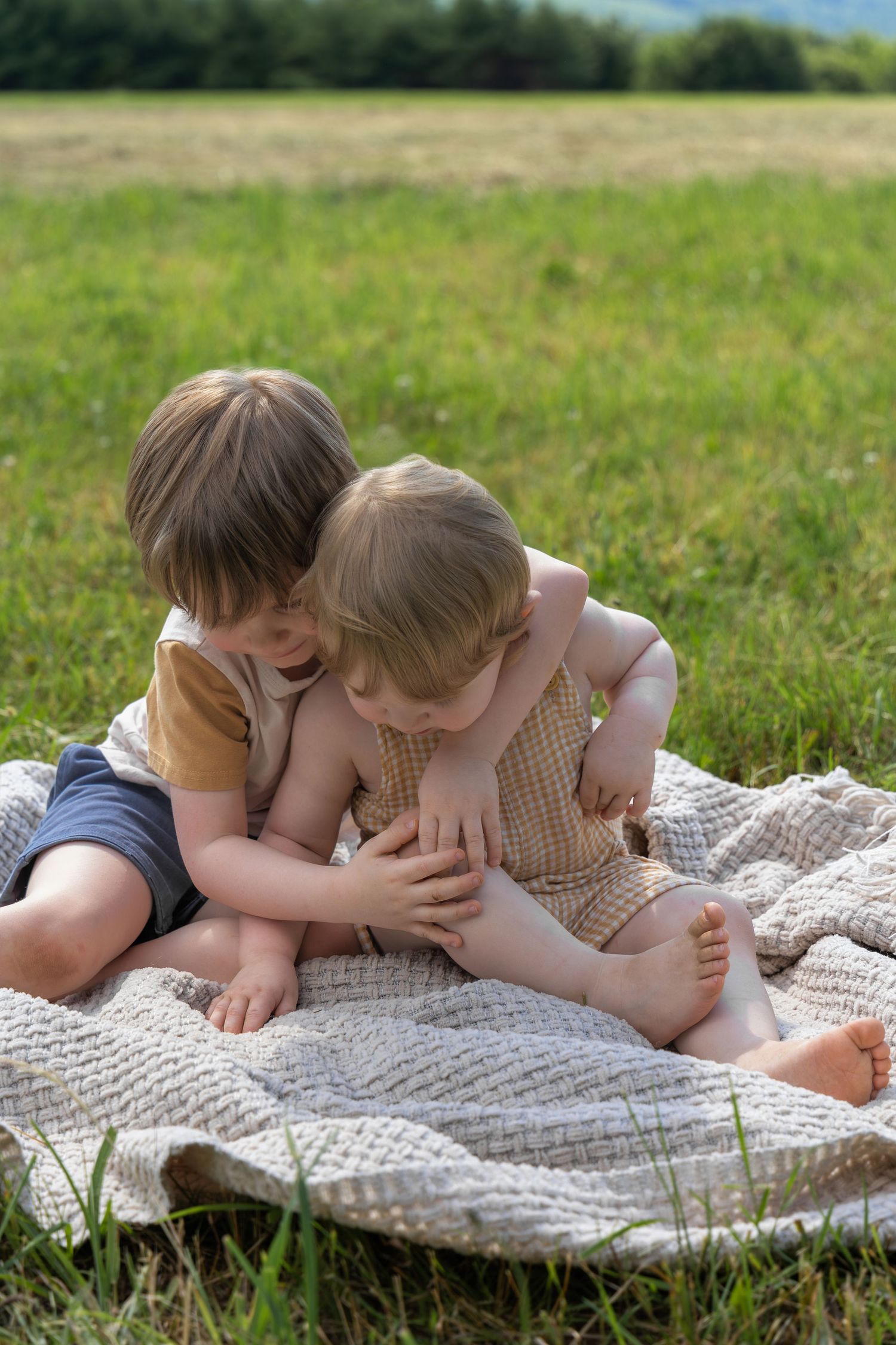 Two young children sitting on a blanket in a green meadow, sharing a sweet sibling moment outdoors.