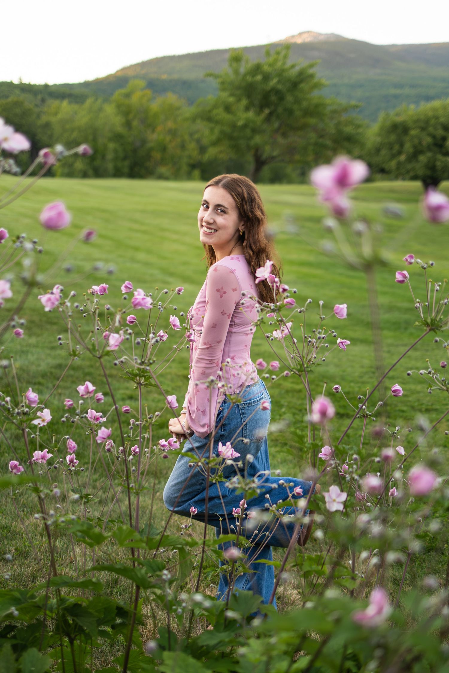 Young woman in pink sweater and jeans smiling among pink wildflowers in a lush green meadow with mountains behind.