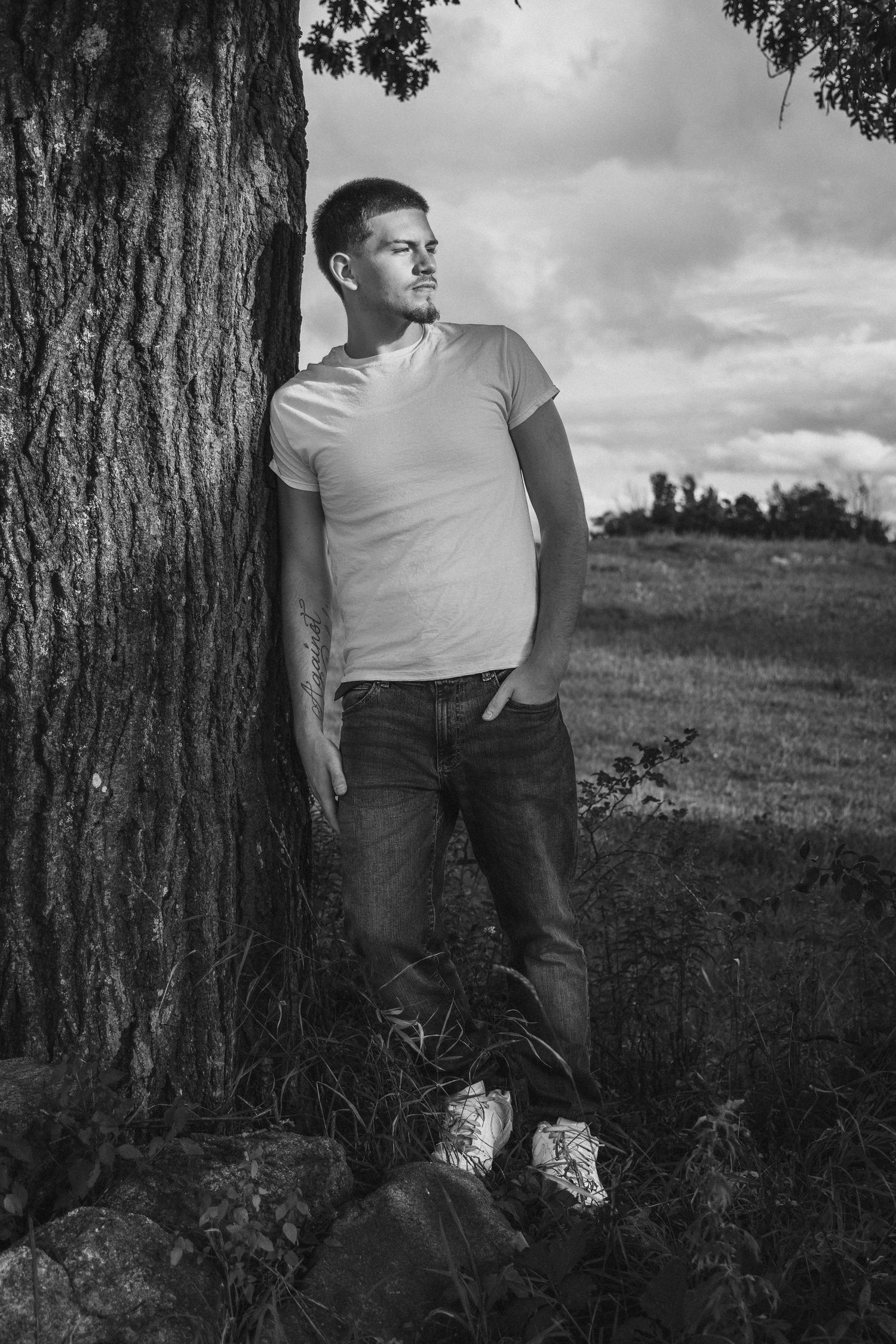 Black and white photo of a young man leaning against a tree in jeans and t-shirt, gazing into the distance outdoors.