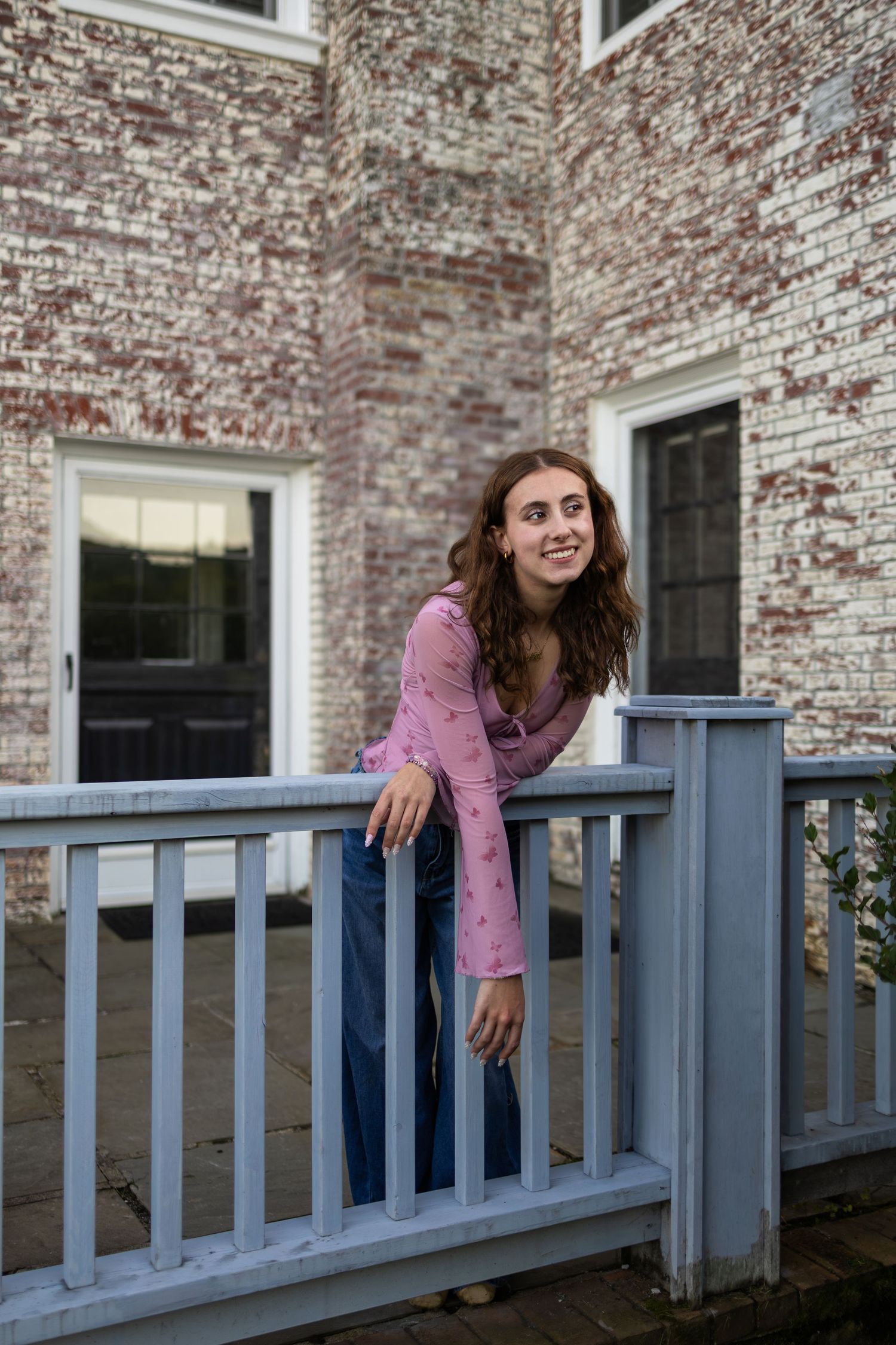 Young woman in pink top and jeans leaning on a blue porch railing in front of a brick building.