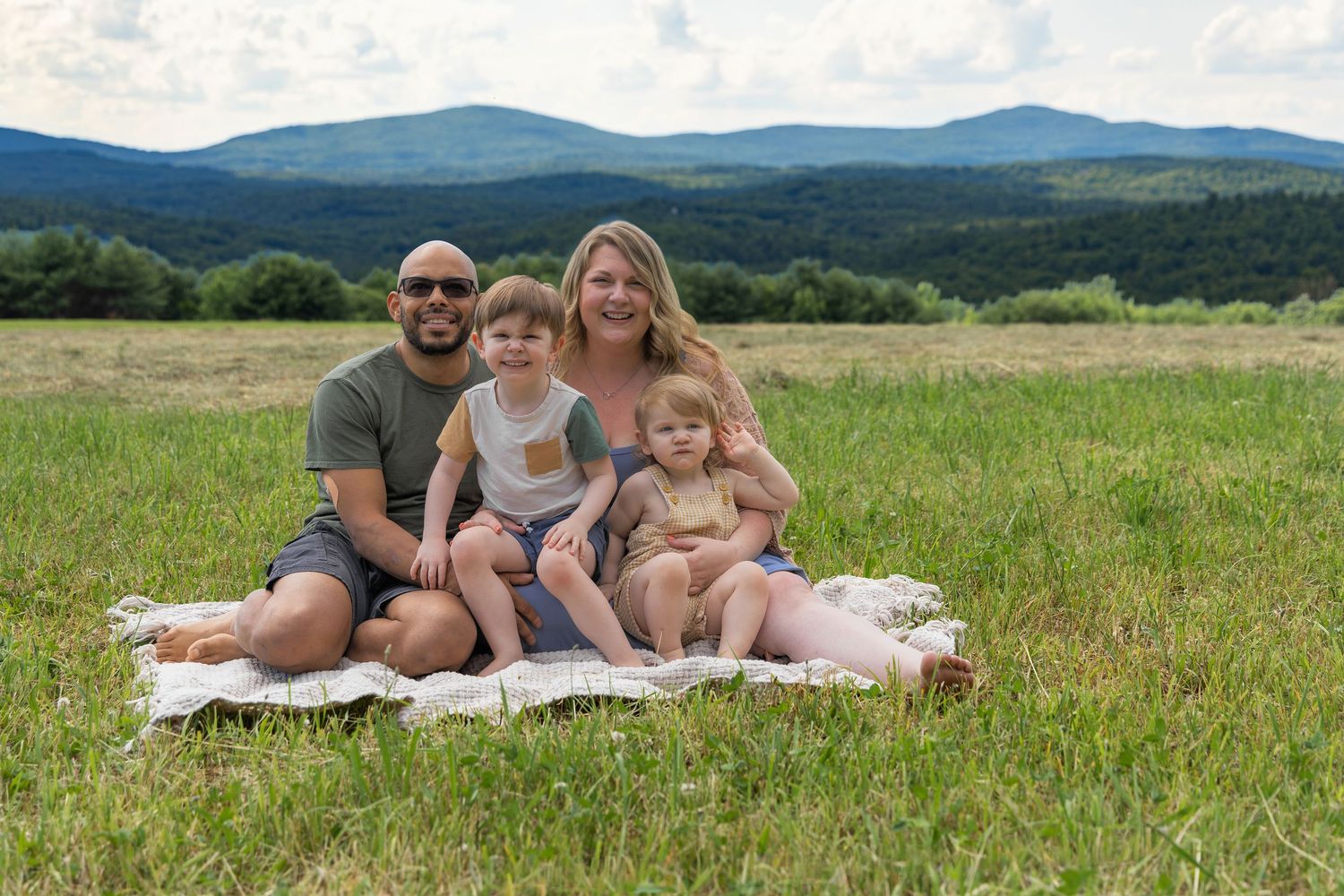 Family of four sitting on a blanket in a green meadow with mountain views on a sunny summer day.