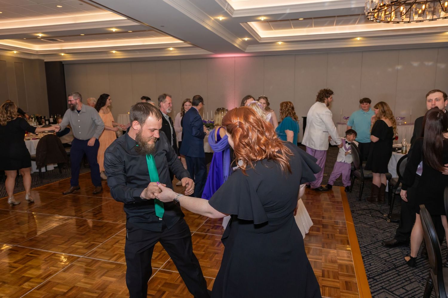 Wedding guests dancing and celebrating in a dimly lit ballroom with wooden floors.