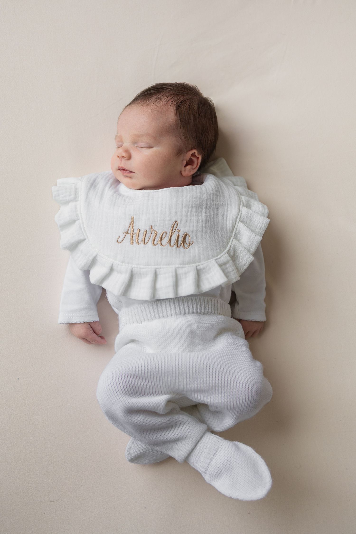 Newborn baby Aurelio sleeping peacefully in white knit outfit with personalized ruffle bib on cream background.
