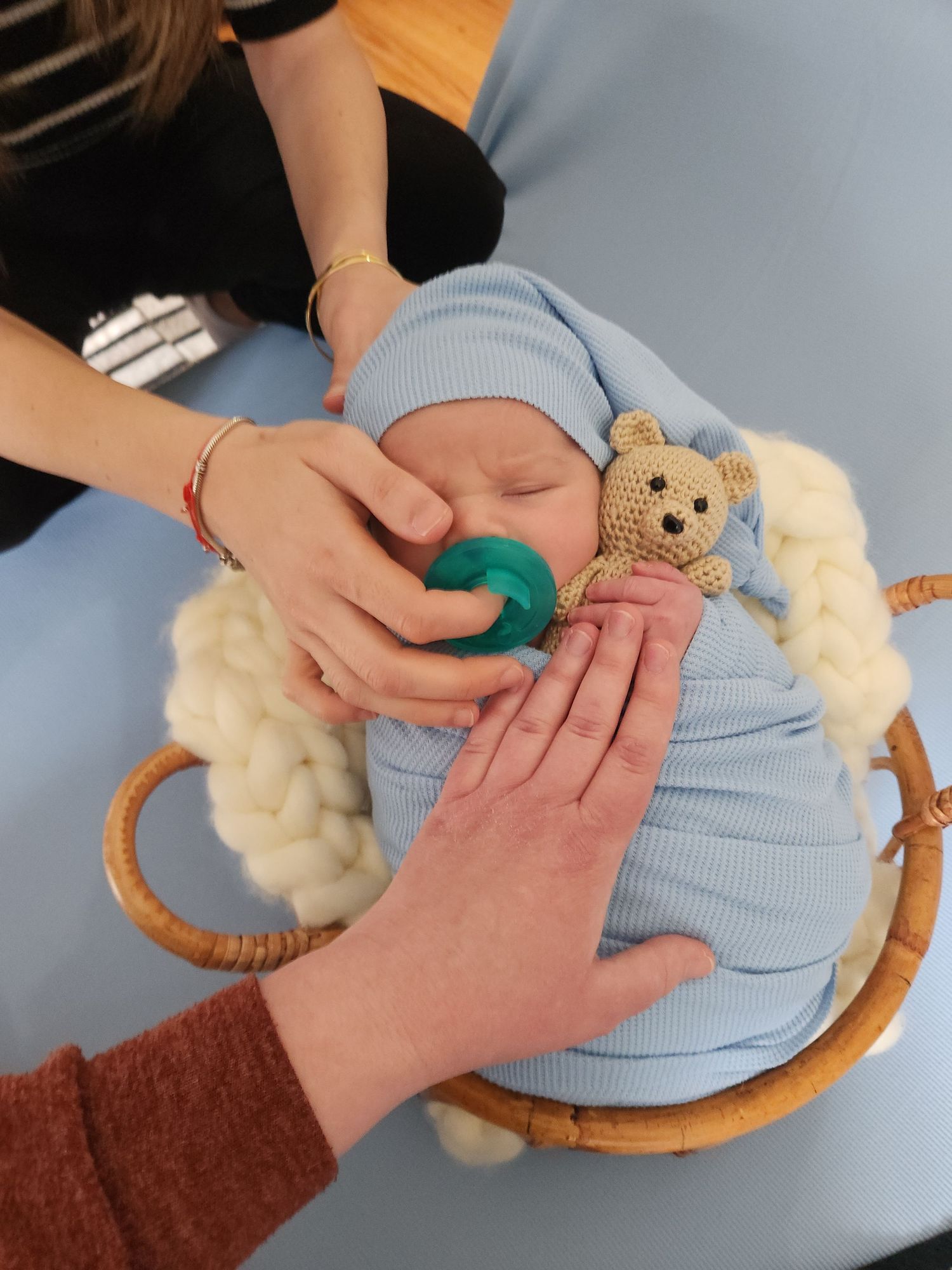 Newborn baby in blue swaddle sleeping in wicker basket with teddy bear, held by two adults.