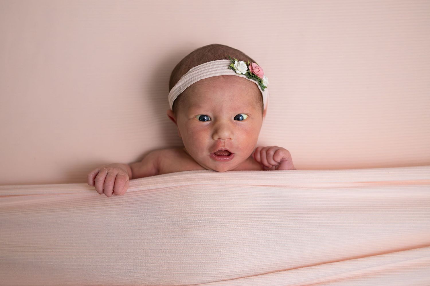 Newborn baby girl wearing a floral headband peeks over a pink blanket in a soft studio portrait.
