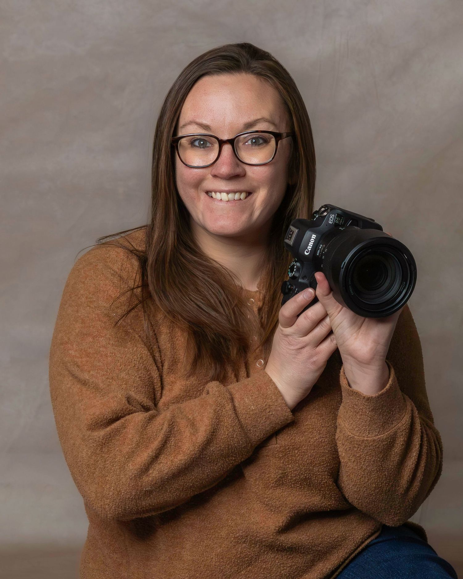 Professional photographer in brown sweater posing with camera against gray backdrop.