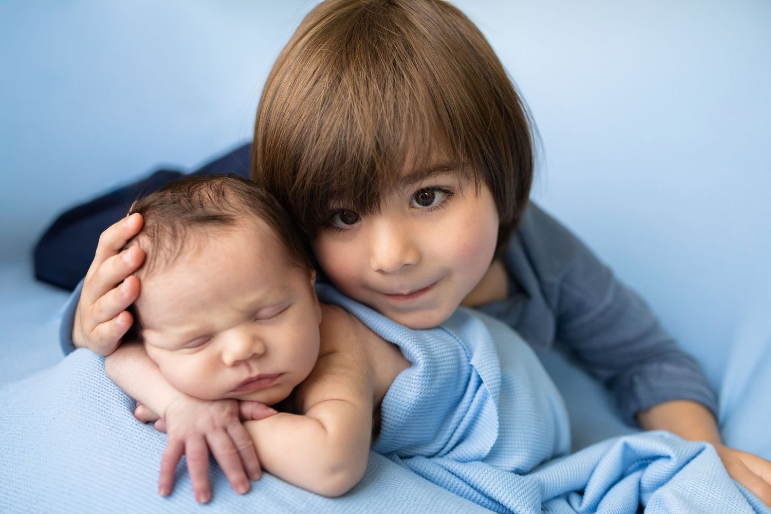 Older child gently holding sleeping newborn baby wrapped in blue blanket, sibling love portrait.