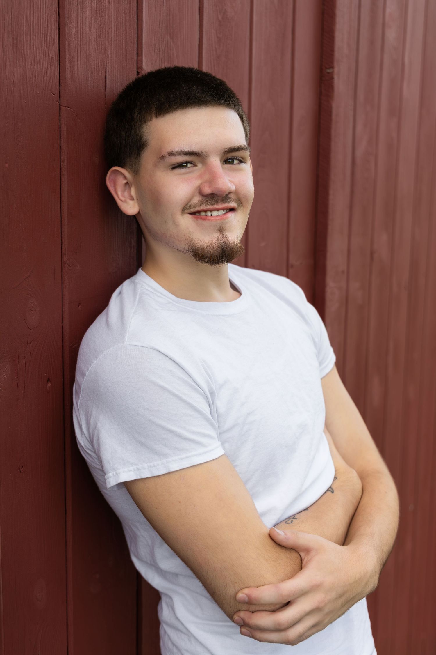Young man with goatee in white t-shirt smiling with arms crossed against a rustic red wooden wall.