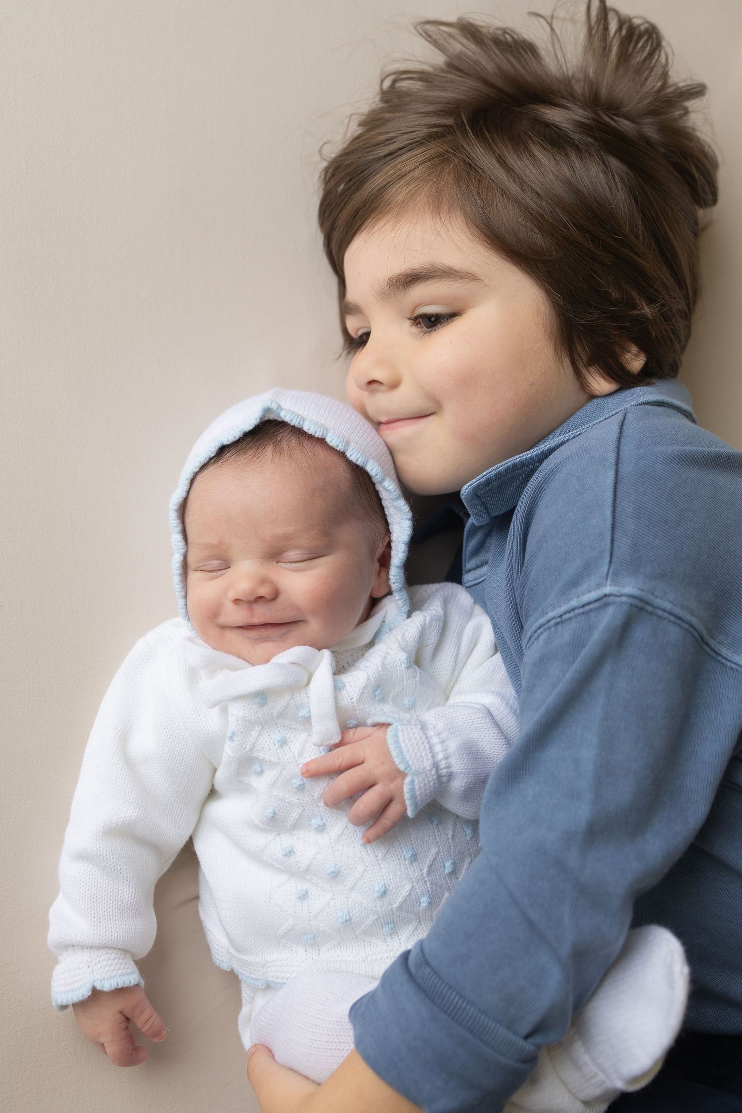 Young boy in blue hoodie gently holding smiling newborn baby dressed in white outfit and bonnet.