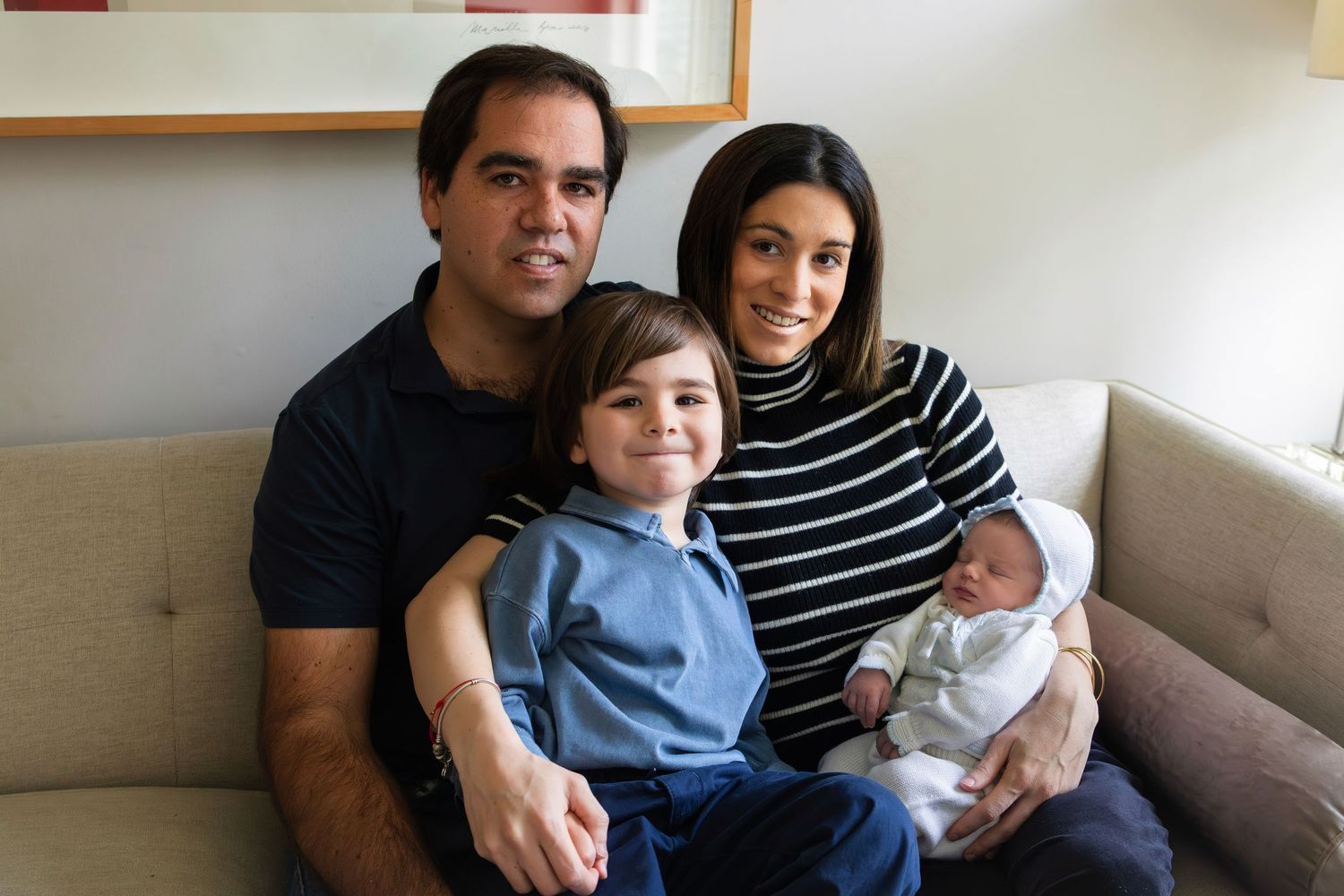 Happy family of four posing on a couch, parents with young boy and newborn baby in a bright living room.
