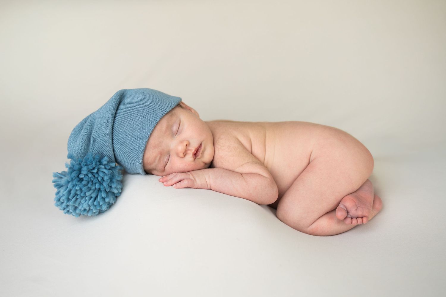 Sleeping newborn baby wearing a blue knit pom-pom hat, curled up peacefully on a white background.