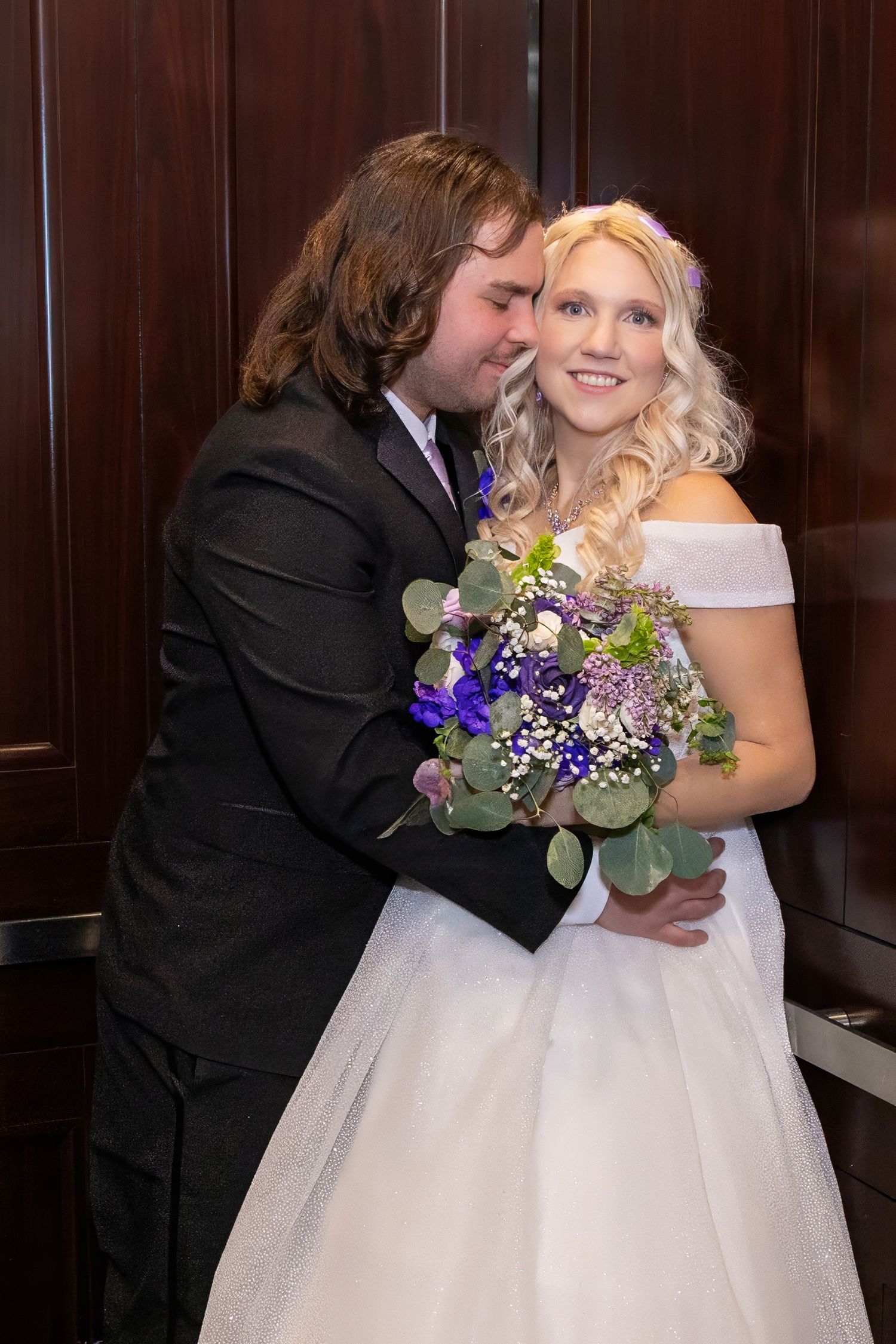 A couple shares an intimate moment in front of dark wooden paneled walls with purple floral bouquets.