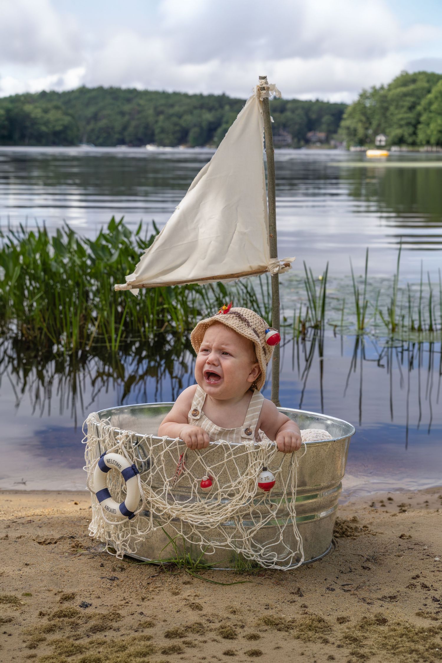Crying baby in sailor hat sits in metal tub with toy sail and fishing net on sandy lake shore.
