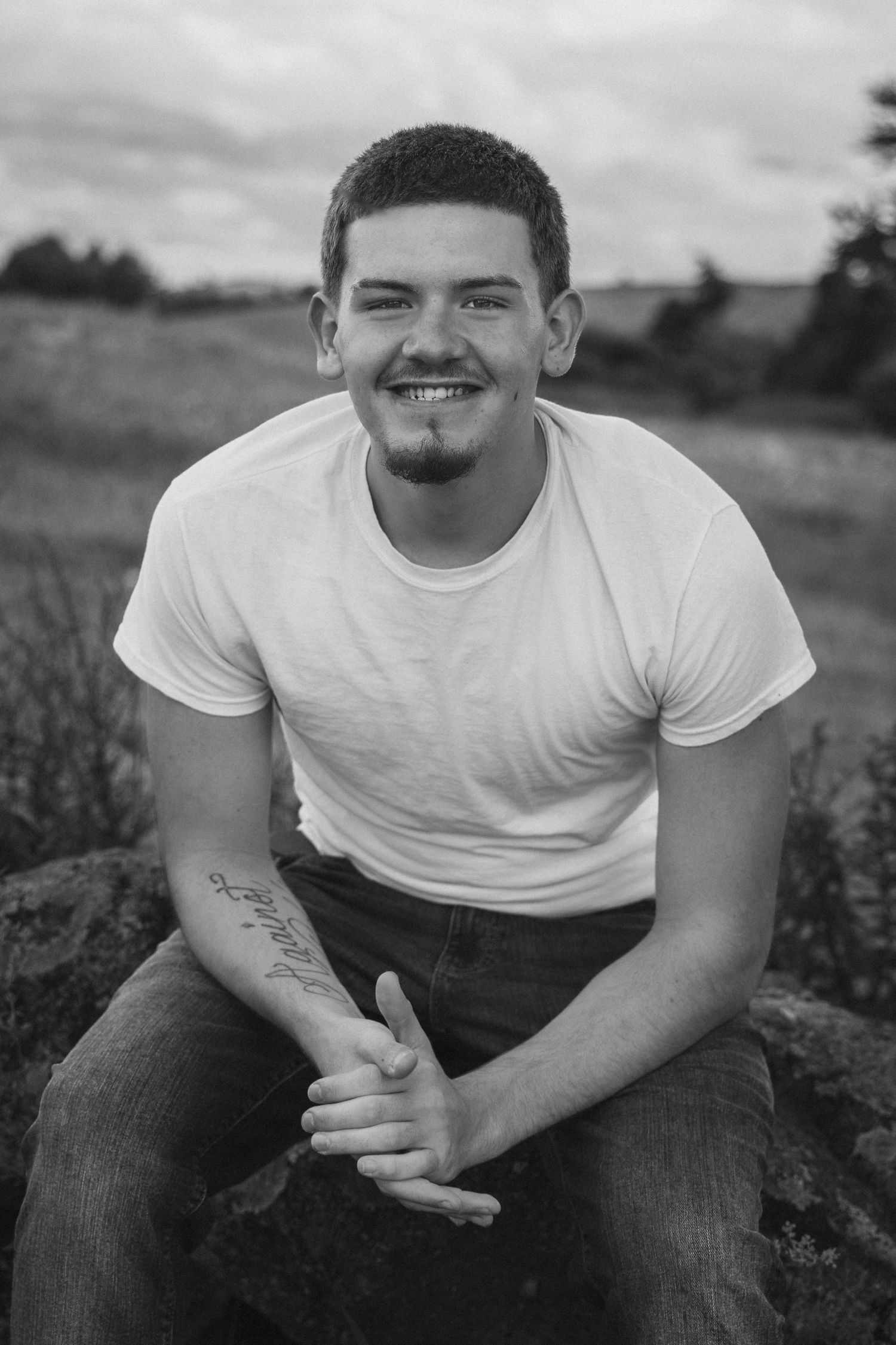 Young man with tattoo smiling outdoors, sitting on rocks in a white t-shirt and jeans, black and white portrait photo.