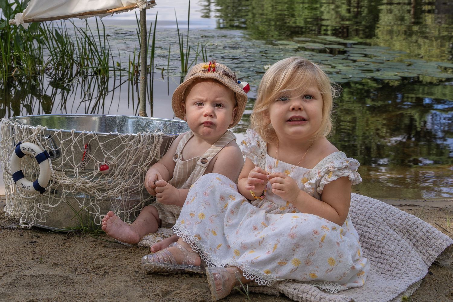 Two toddler girls sitting by a lake shore with a fishing net basket, wearing summer dresses on a sunny day.