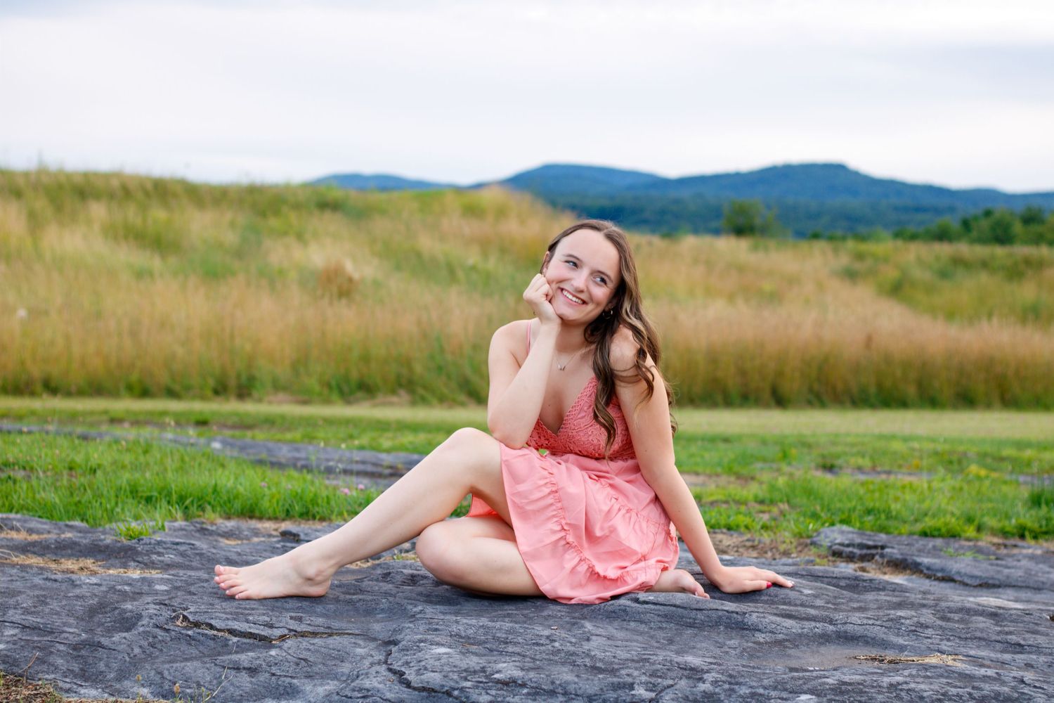A person in a pink dress poses on a large rock formation with mountains in the background.
