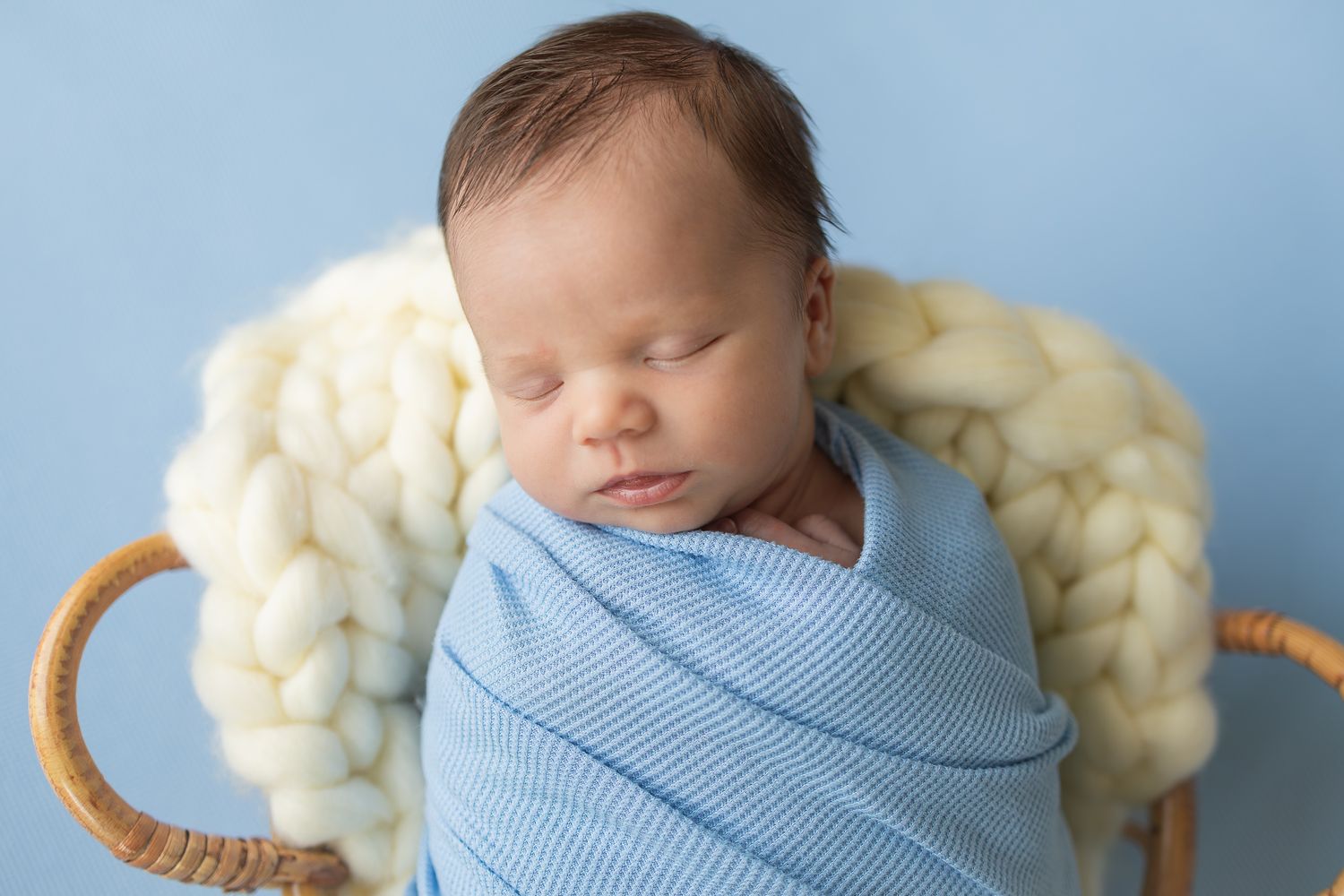 Newborn baby swaddled in blue wrap sleeping peacefully in a wicker basket with cream chunky knit blanket.