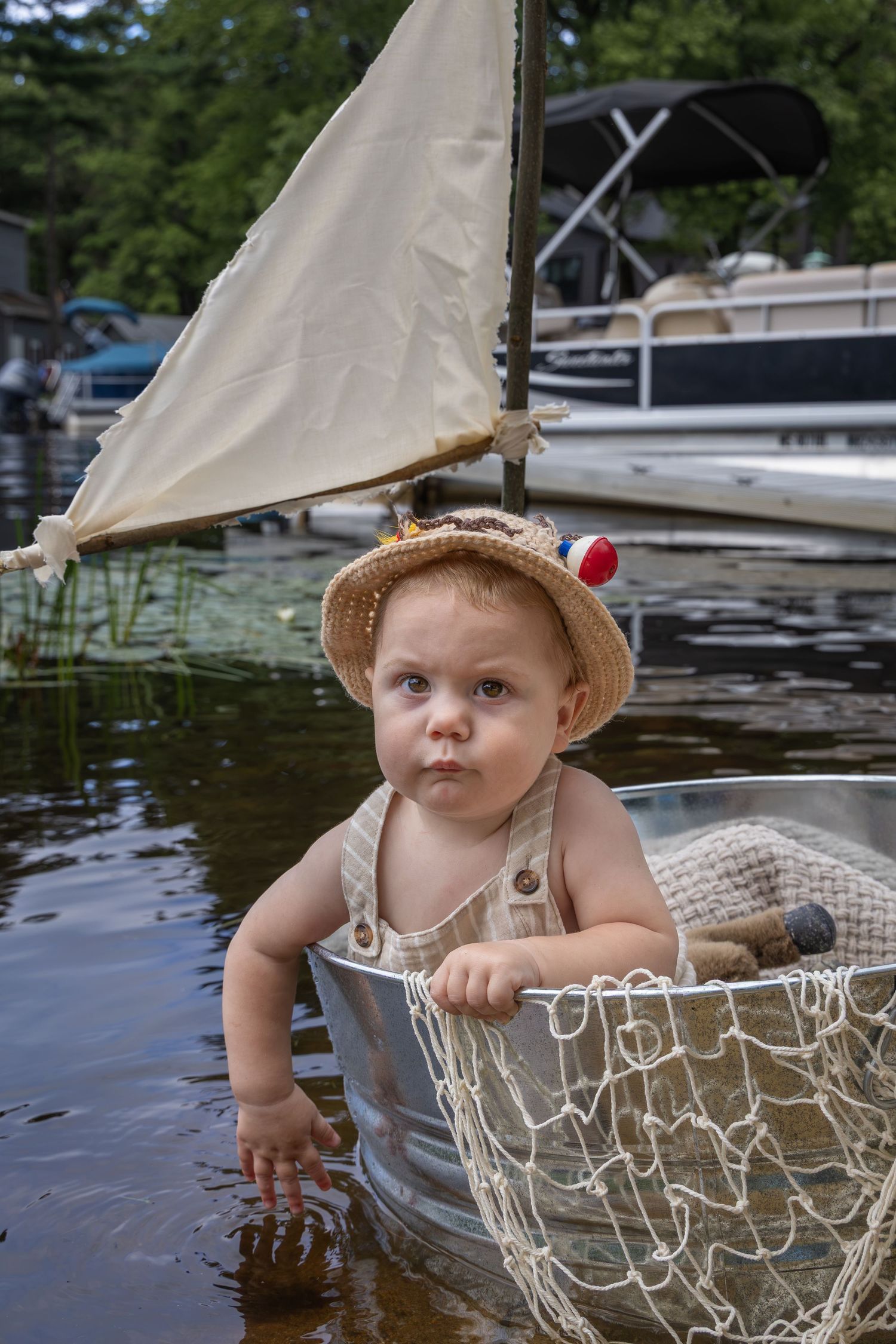 Adorable baby in straw hat sits in small metal boat with fishing net on calm lake, sailboat visible behind.