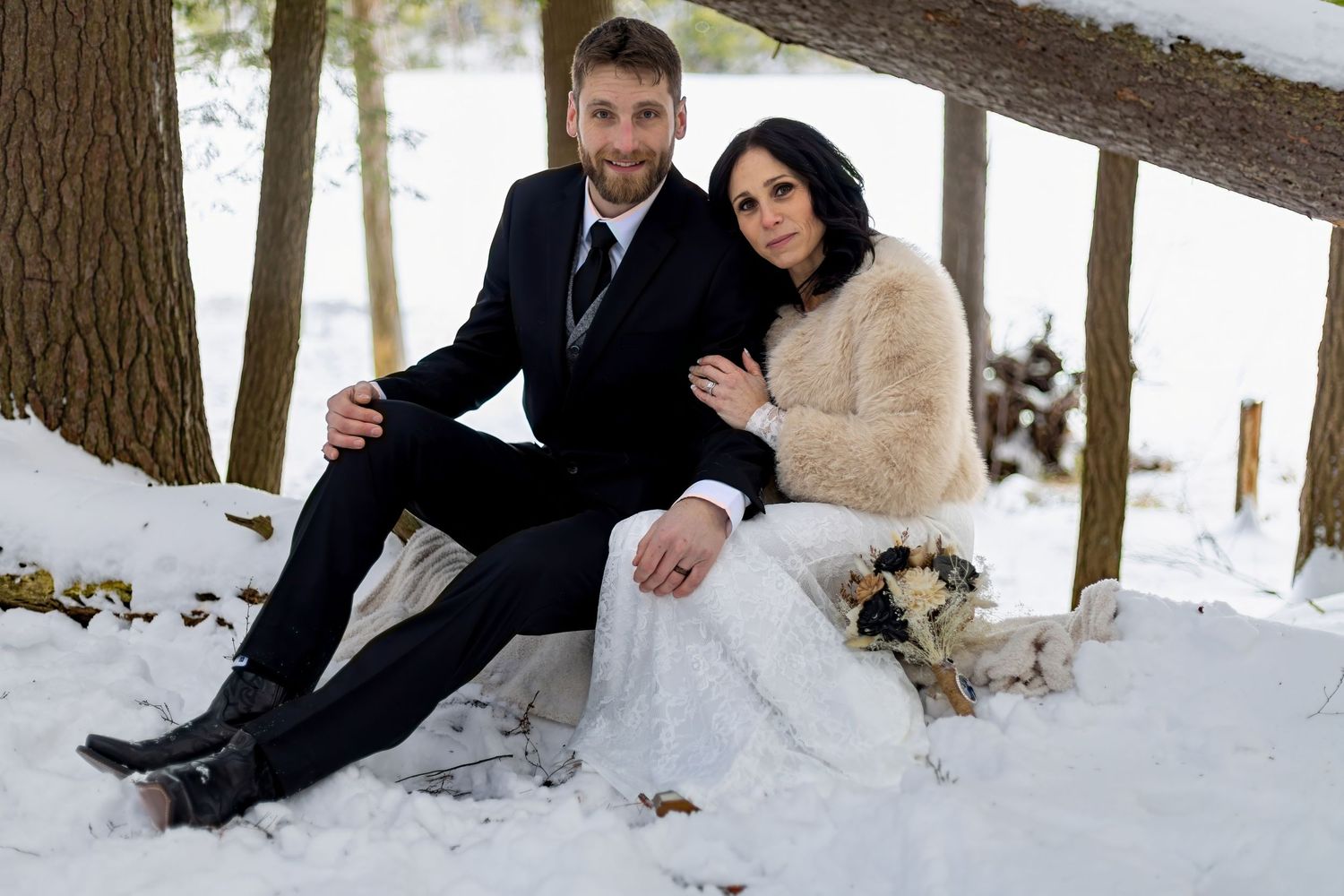 Wedding couple posing playfully in the snow near tree trunks.