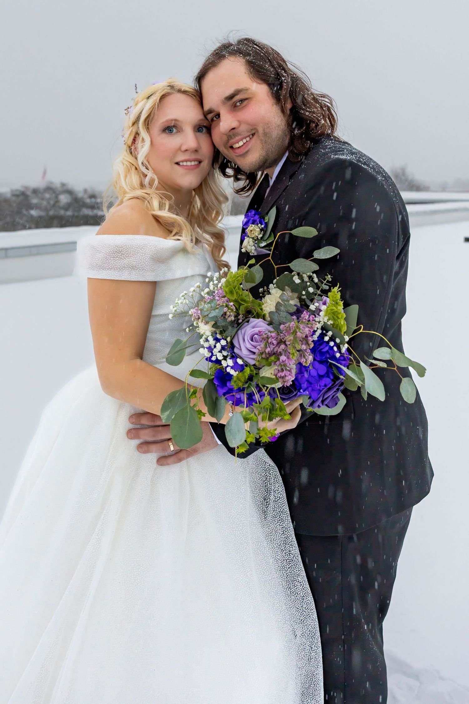 Romantic winter wedding portraits with snow falling and purple floral accents.