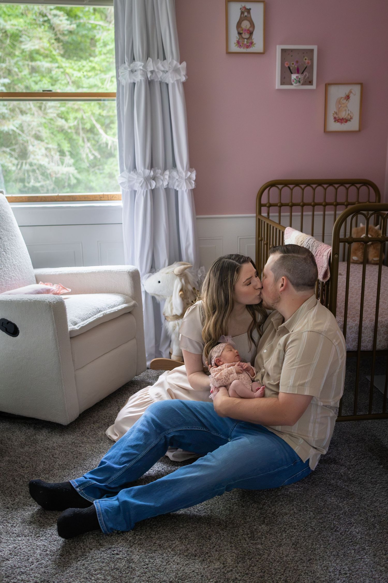 New parents sitting on nursery floor kissing while holding their newborn baby in a pink decorated room.