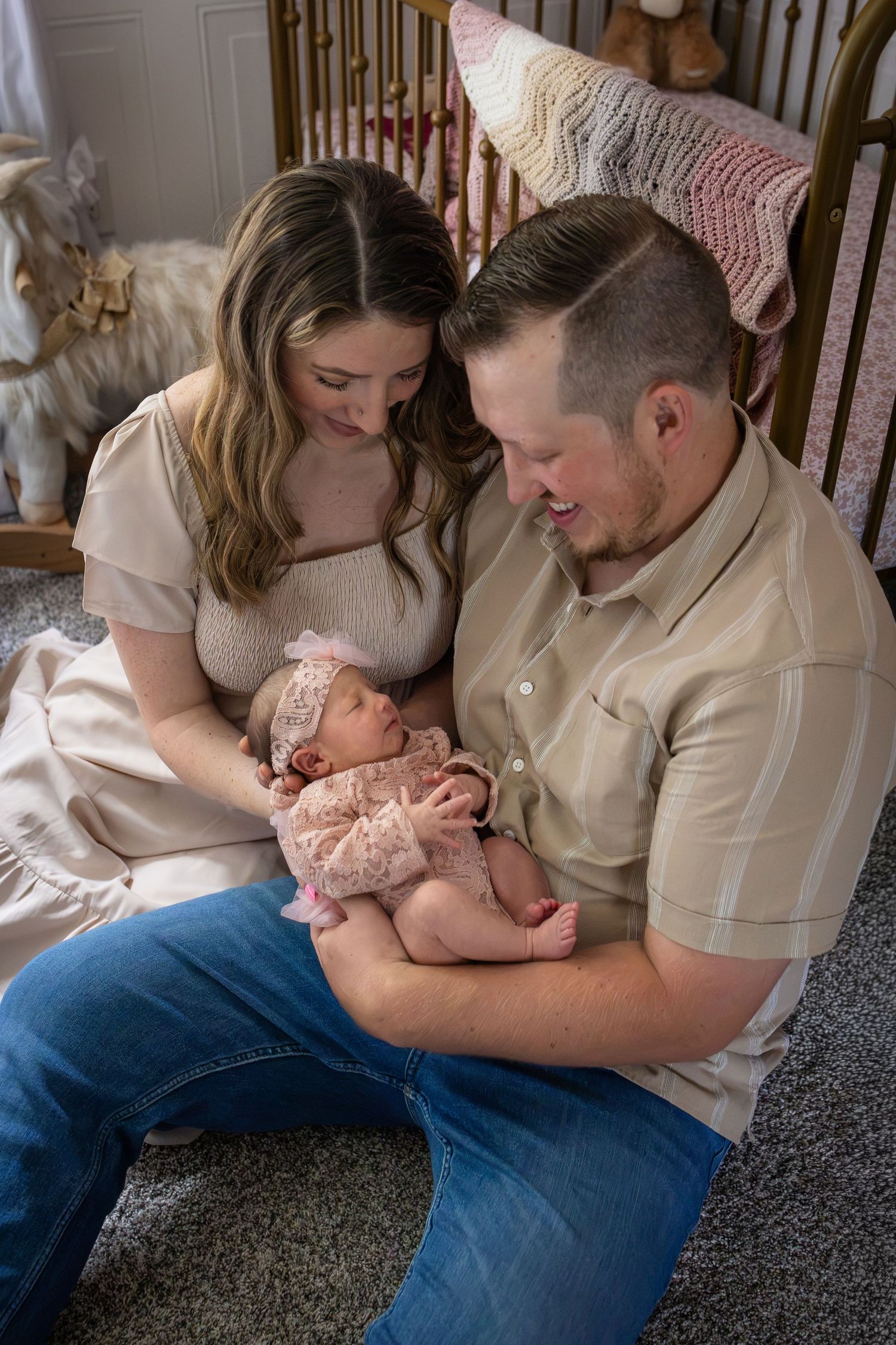 New parents sit on nursery floor smiling at their newborn baby girl dressed in pink, creating a tender family moment.