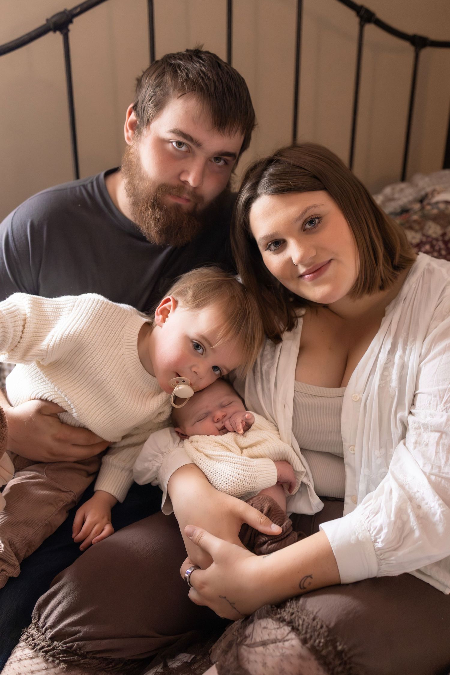 A young family poses together on a bed, parents smiling with a toddler and newborn baby in a warm, cozy setting.