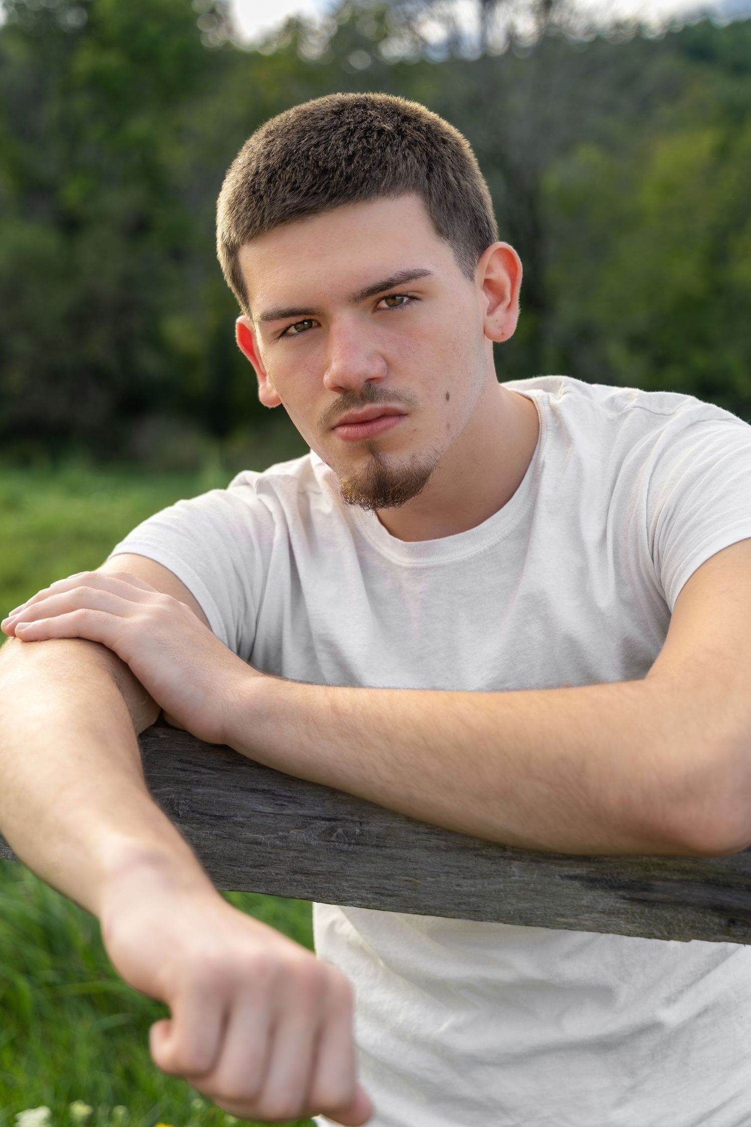 Young man in white t-shirt leaning on wooden fence outdoors, with green trees in background.
