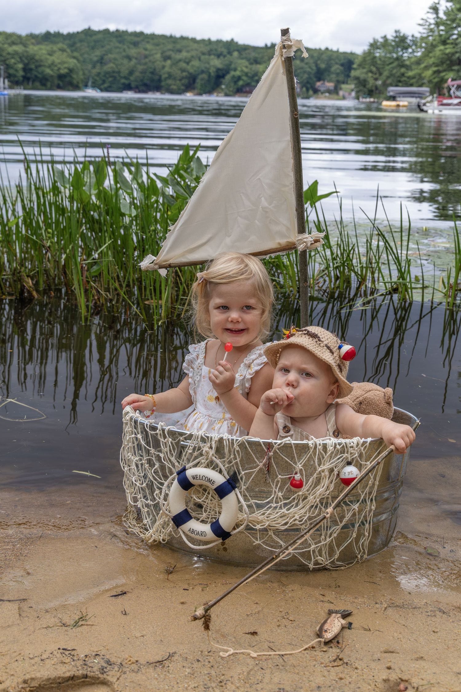 Two toddlers sitting in a decorative boat basket with a sail on a sandy lakeshore.