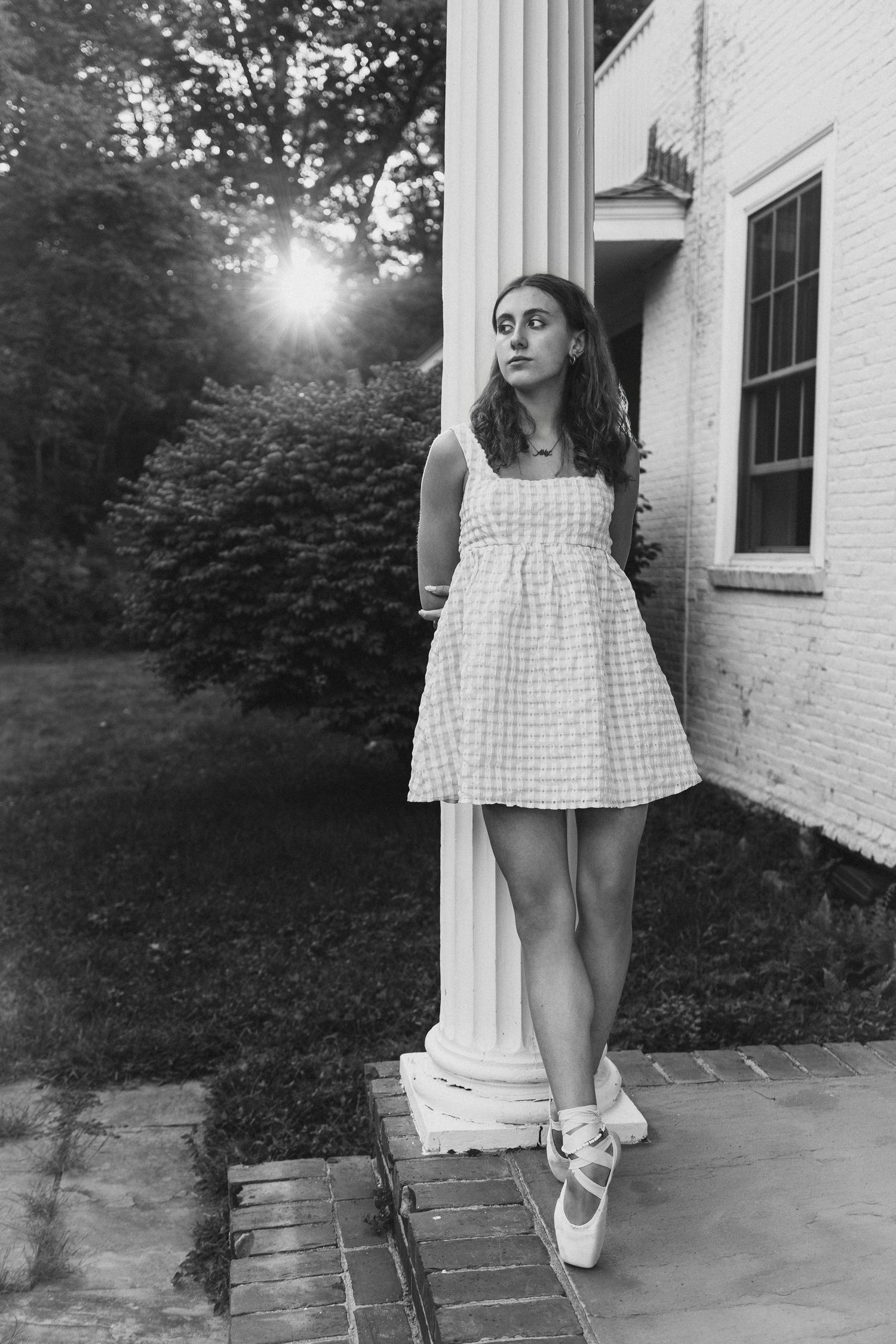 Black and white photo of a young woman in a gingham dress leaning against a white column at golden hour.