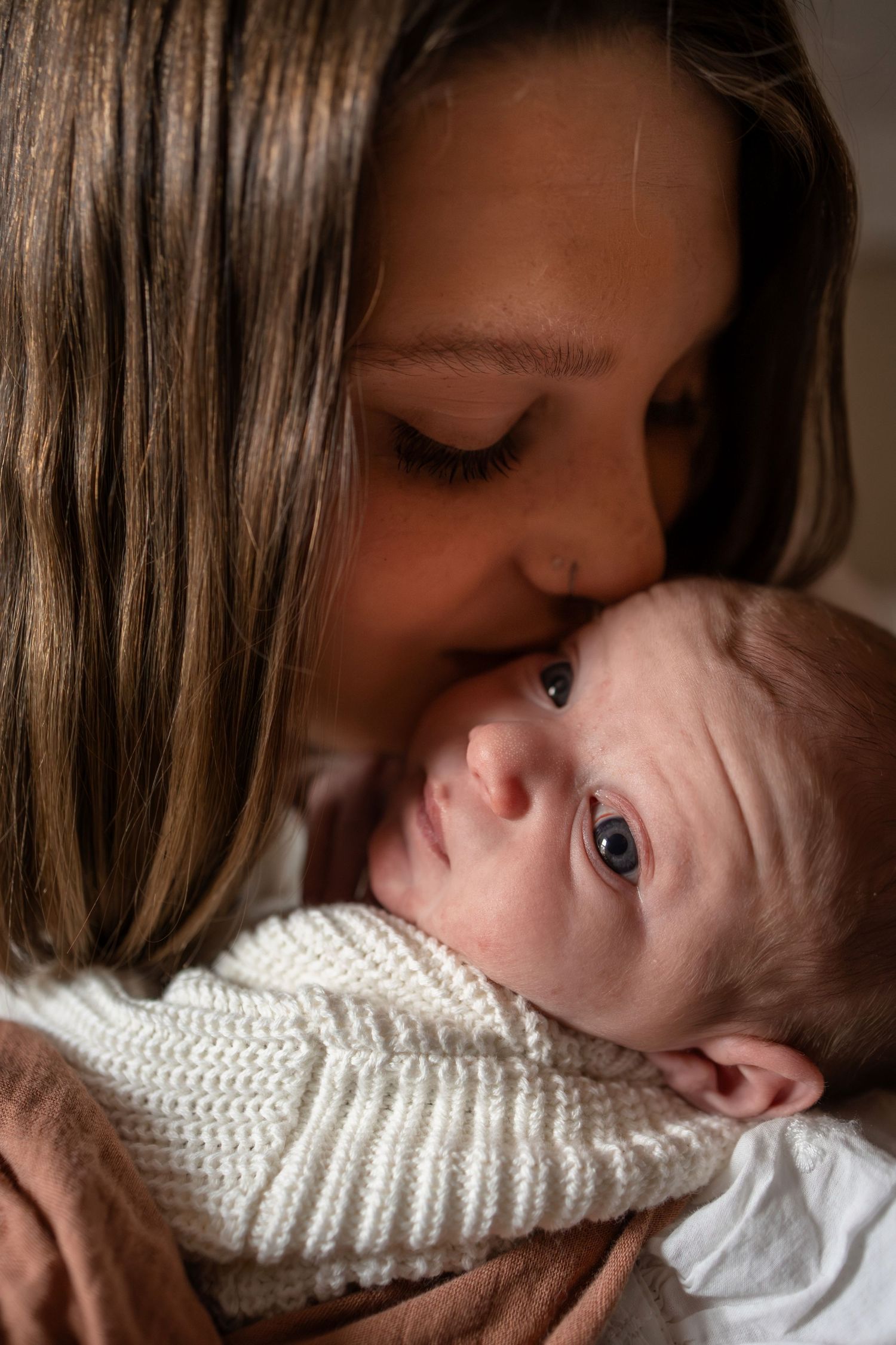A tender mother gently kissing her newborn baby wrapped in a white knit blanket.