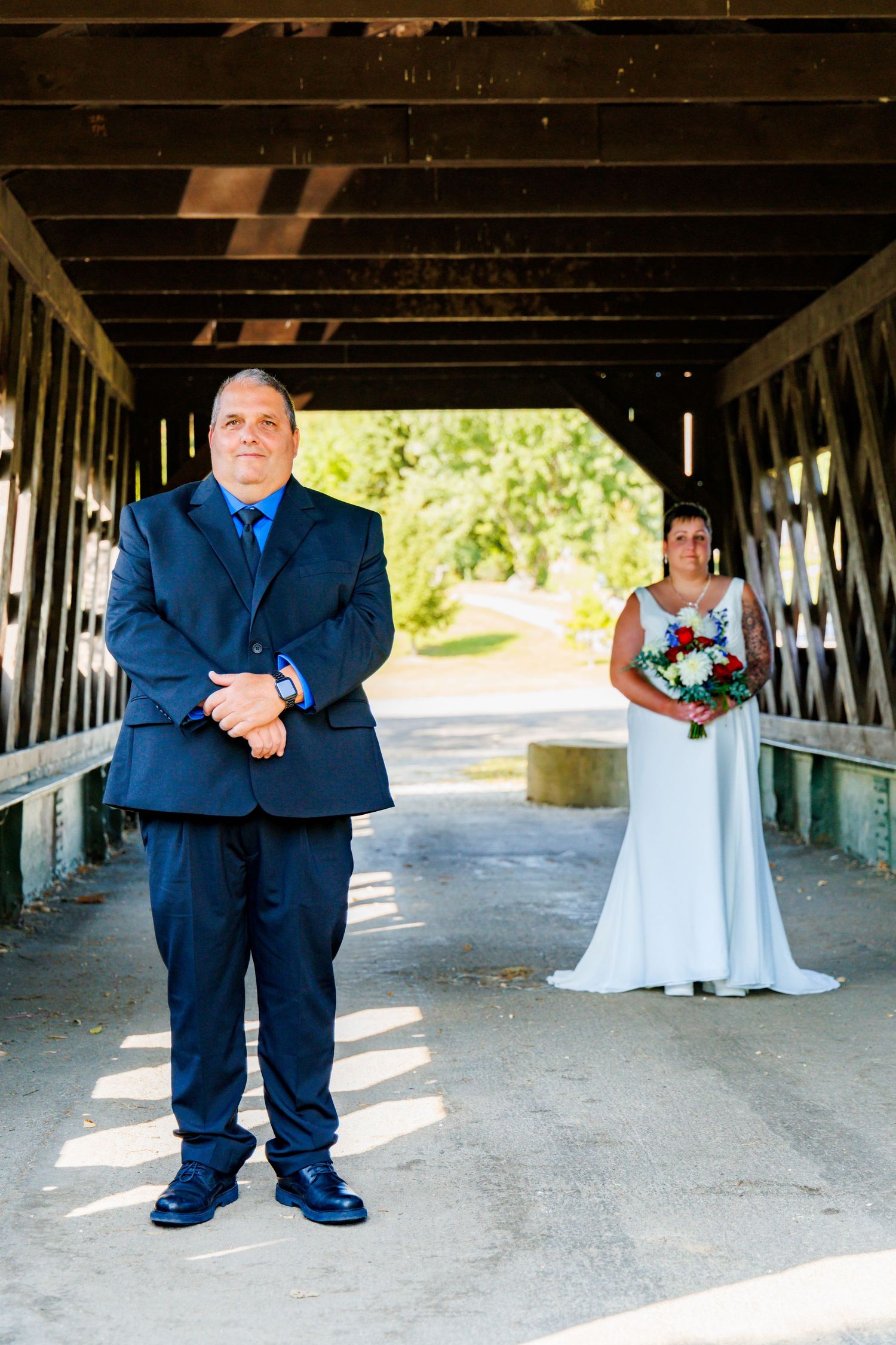 Wedding party members in navy suits and white dress stand under a rustic covered bridge.